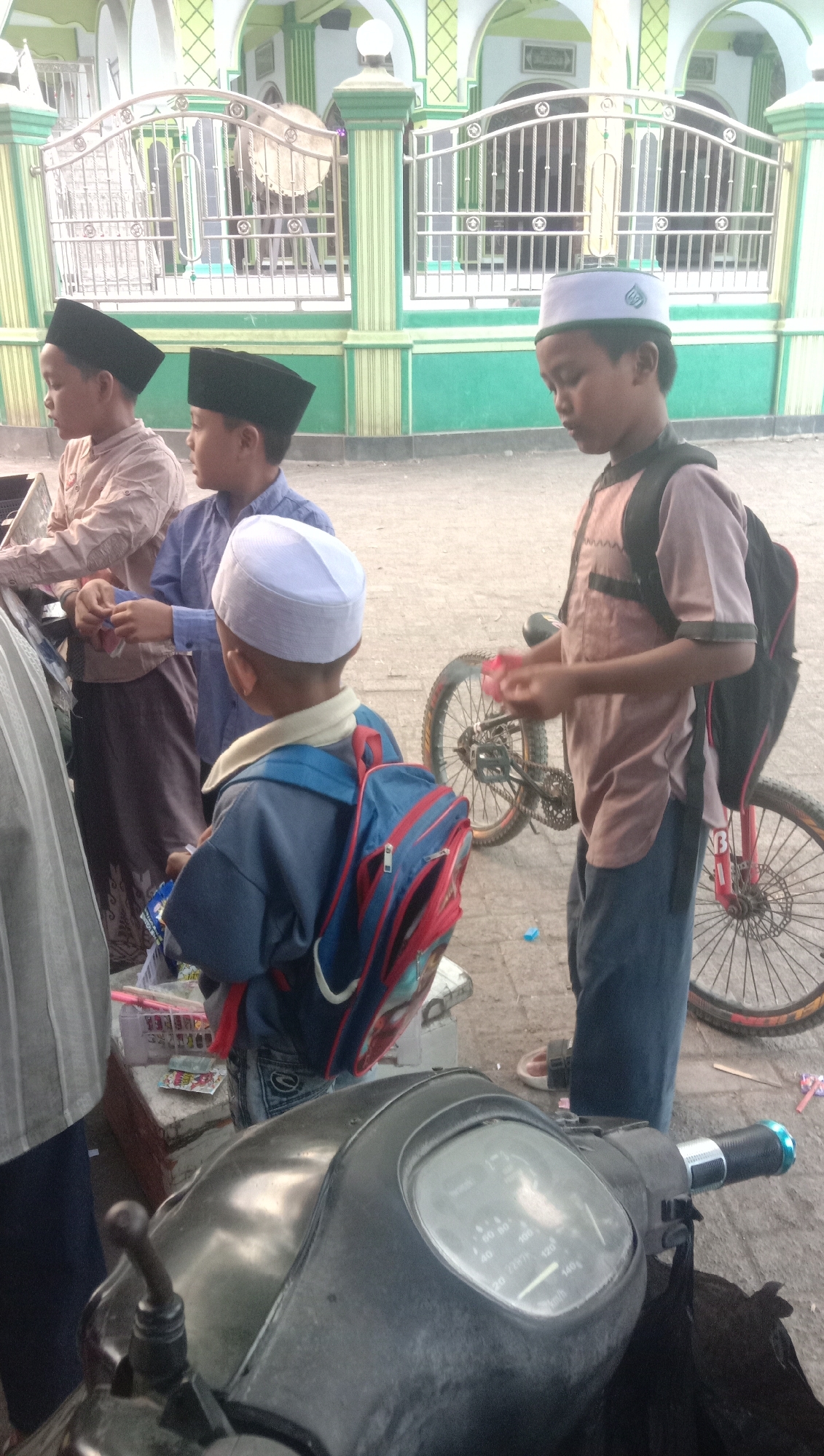 In Ngempit, Indonesia, four boys are gathered near a building with green and white architectural elements, possibly a mosque. The building features a metal fence with vertical bars and decorative circular elements. Two boys in the foreground wear skullcaps, one black and one white. The boy with the white skullcap is facing away from the camera and has a blue jacket with a red and blue backpack. The boy with the black skullcap, positioned behind him and to the left, wears a light brown shirt and a black skullcap, and appears to be holding something in his hands. Further to the left, another boy in a light brown shirt and black skullcap is visible, holding an object. A fourth boy, standing further back on the right, wears a light brown shirt, dark pants, and a black backpack. He is holding a pink object and standing next to a bicycle with red accents on the wheels. In the immediate foreground, the handlebars and dashboard of a scooter are visible. The ground is paved with asphalt.