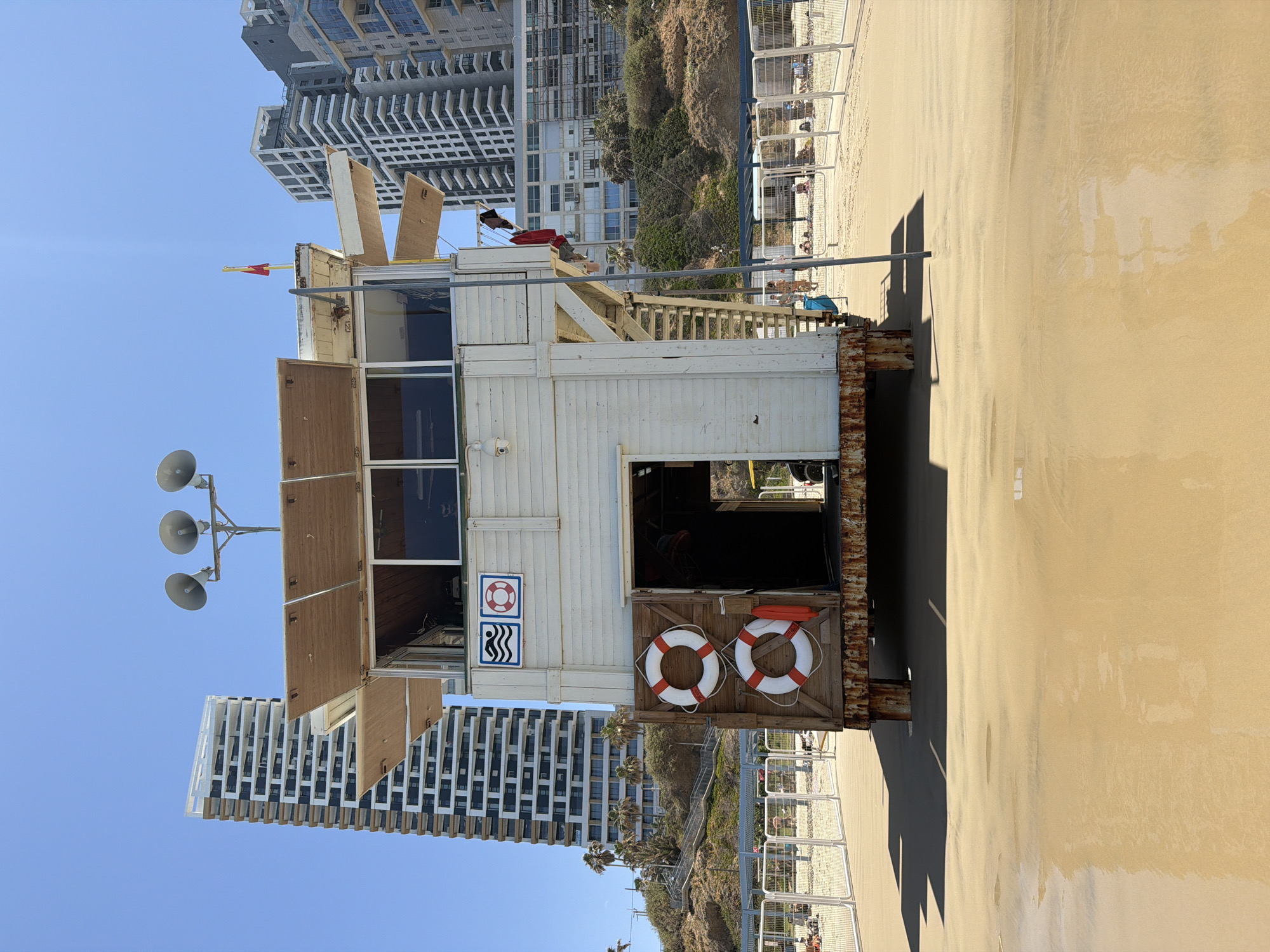 The image shows a lifeguard station on a beach. The station is made of wood and elevated on stilts, with two lifebuoys hanging on its front. The station has an open entrance with the inside unseen. On top of the station are three loudspeakers. In the background are modern high-rise buildings of Bat Yam, Israel, and a path leading up to the dunes next to the beach. The sand is light brown, and the sky is blue. The station is casting a shadow on the sand. A fence is present behind the station, separating the dunes from the beach. There are people on the beach beyond the fence.