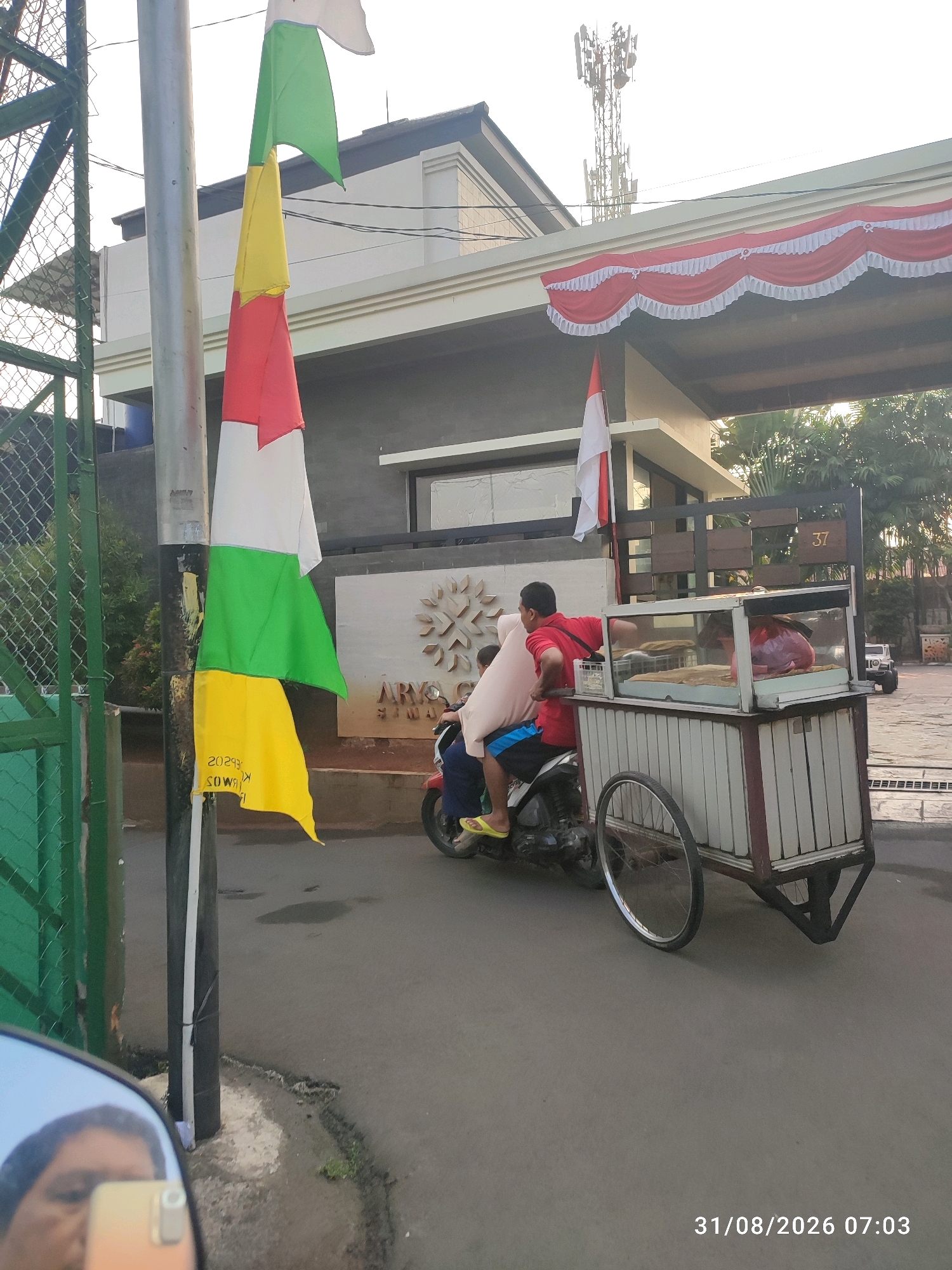 The image shows a street scene in East Jakarta, Indonesia. In the foreground, a motorcycle with a sidecar is parked. The sidecar is a mobile food cart with a glass display case, containing food items. A person wearing a red shirt and blue shorts is seated on the motorcycle, holding onto the cart. Another person is partially visible behind them. A tri-colored flag, red, white, and green, is attached to a pole to the left of the motorcycle. The flag has some writing on it. In the background, there is a building with a red and white awning. A cell tower is visible in the distance. The road surface is asphalt. The timestamp "31/08/2026 07:03" is present in the bottom right corner of the image.