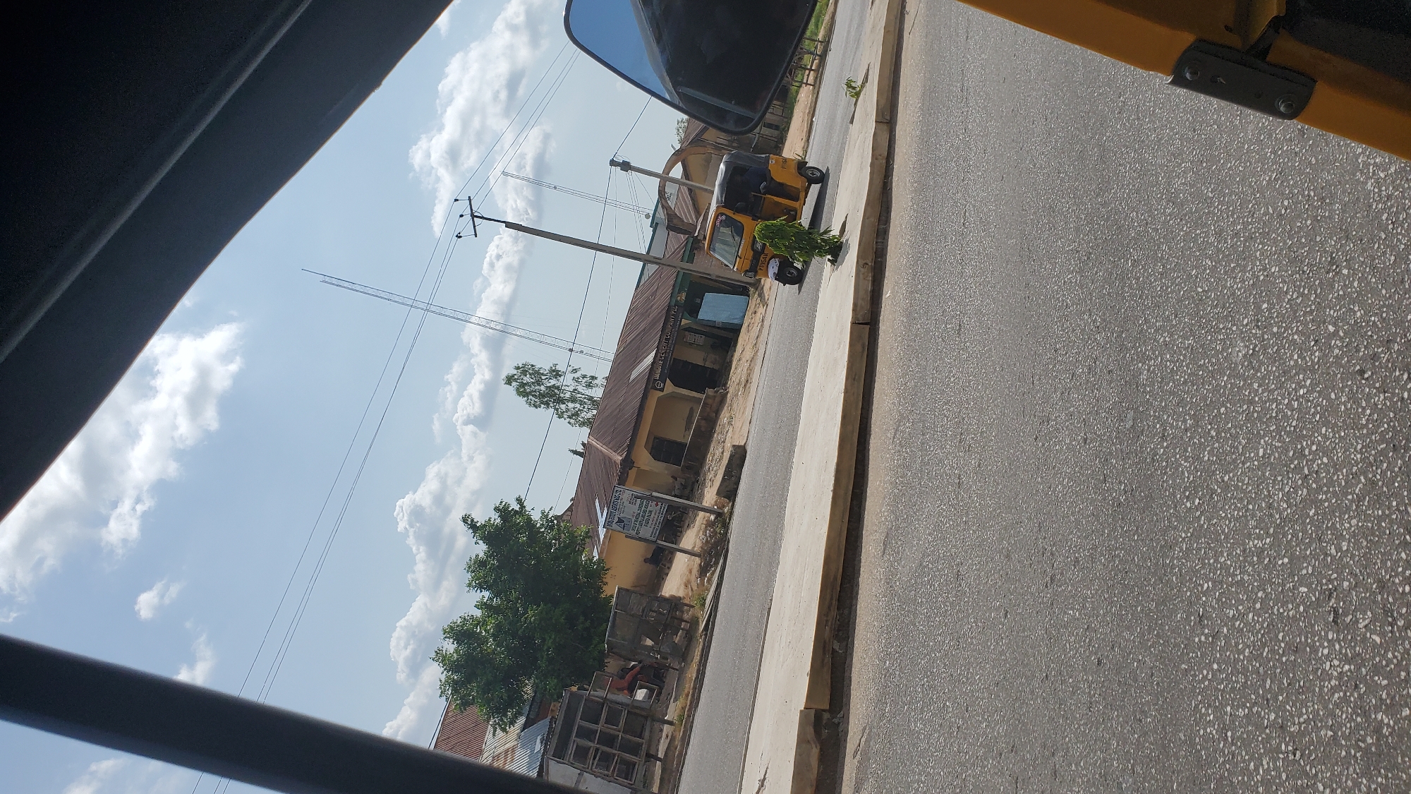 The image captures a street scene in Jalingo, Nigeria, under a partially cloudy sky. In the foreground, a yellow motorized tricycle, commonly known as an "Aja" or "Keke," is parked on the right side of the road, facing away from the viewer. The tricycle has a black roof and a driver is visible inside. A small green bush is planted in front of the tricycle.
The road itself is paved with asphalt and a concrete sidewalk runs along its edge. Beyond the sidewalk, a row of single-story buildings with corrugated iron roofs lines the street. Some of these buildings appear to be shops or small businesses, with one bearing a sign that reads "OKORO MARKETING AGENCY & CO. LTD." Another sign on a different building states "DAKPOL CONCRETE COMPANY PLC." A few people are visible near these buildings, some standing and others seated.
Trees, including a prominent leafy tree on the left, are scattered throughout the scene. Overhead, power lines and a tall communication tower are visible against the sky. The image appears to be taken from a moving vehicle, possibly another tricycle, as indicated by the dark frame on the top left and the partial view of the vehicle's yellow interior on the bottom right. The overall impression is of a typical urban environment in a Nigerian city, with a mix of commercial buildings, transportation, and everyday life.
