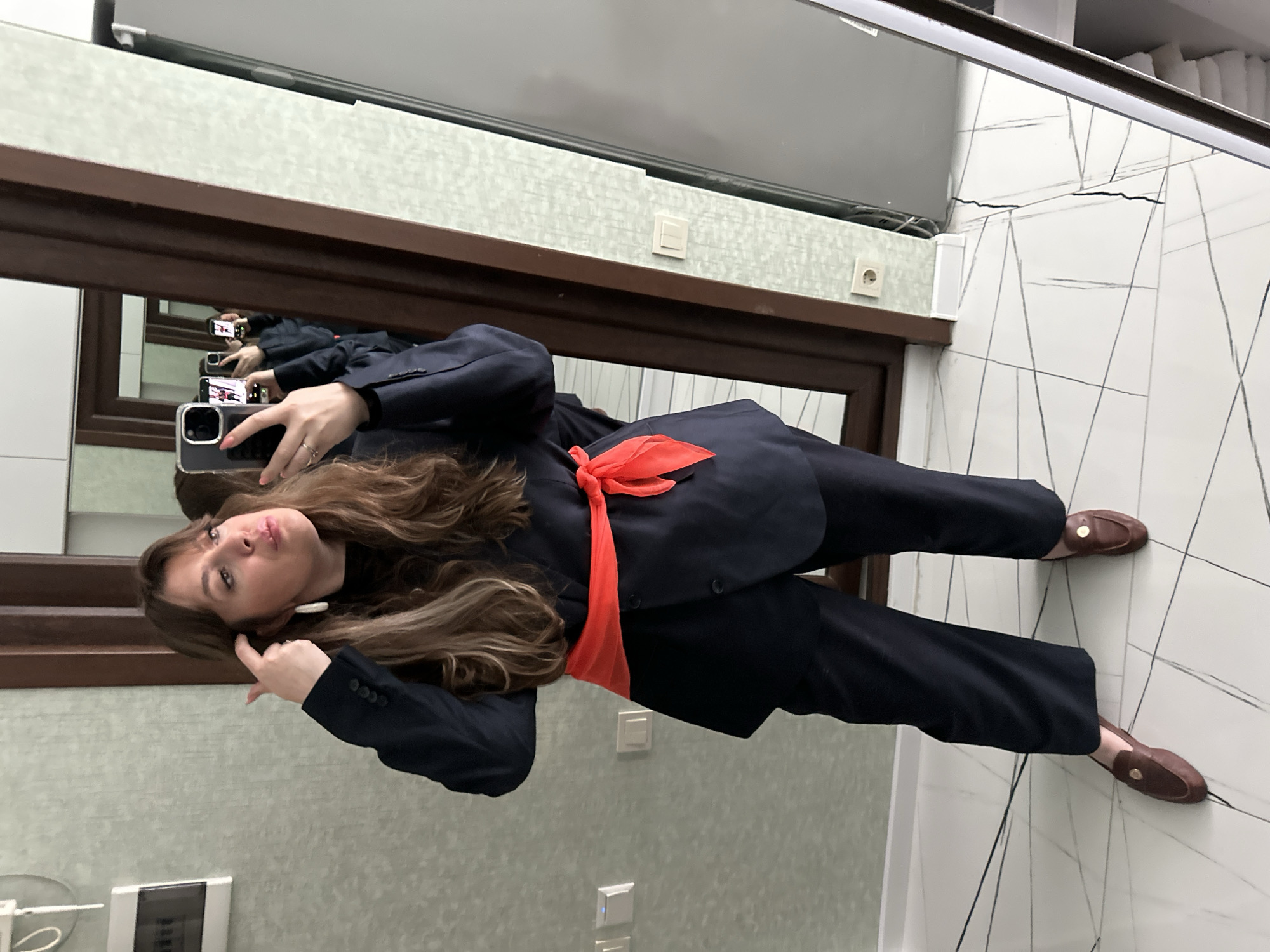 A woman is taking a selfie in what appears to be a bathroom. She is wearing a dark blue pantsuit with an orange scarf and brown loafers. The bathroom has light green walls, a brown-framed mirror, and white tile flooring with black line patterns. The photograph was likely taken indoors in Batumi, Georgia.