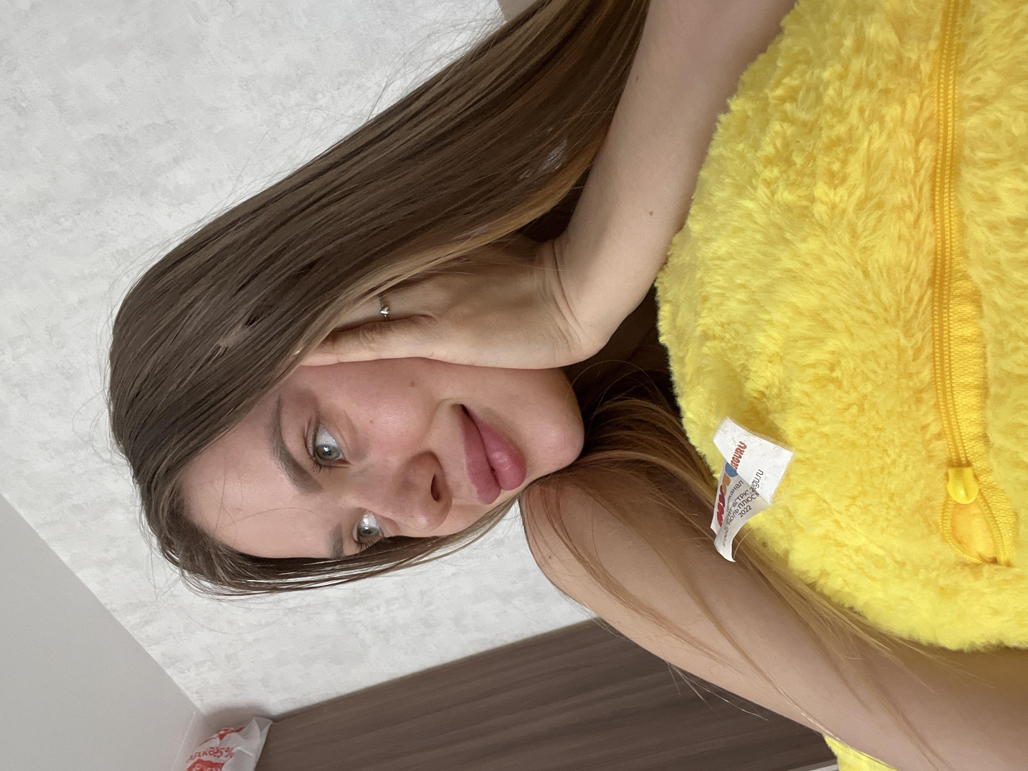 The image, taken in Chaikovsky, Russia, features a young woman indoors. She has long, straight, medium brown hair and light-colored eyes, possibly green or hazel. She is looking directly at the camera with a soft, relaxed smile. Her right hand, adorned with a ring on the ring finger, is gently placed against her face.
She is wearing a vibrant yellow, fluffy garment, resembling a costume or onesie, which features a visible zipper down the front. A white fabric label is attached to the yellow garment, displaying text primarily in Russian. Partially visible words on the label include "GURU," "kanal," "от РКО/РКД," "РУ.RU," "ОНЬ ПЛЮС," and the year "2022."
The background suggests an indoor setting, with a light-colored wall or ceiling on one side and a darker, possibly wooden, surface on the other. A small portion of a white plastic bag is visible in the lower-left corner. The image is rotated 90 degrees clockwise from its likely original orientation. The overall impression is that of a casual and pleasant moment.