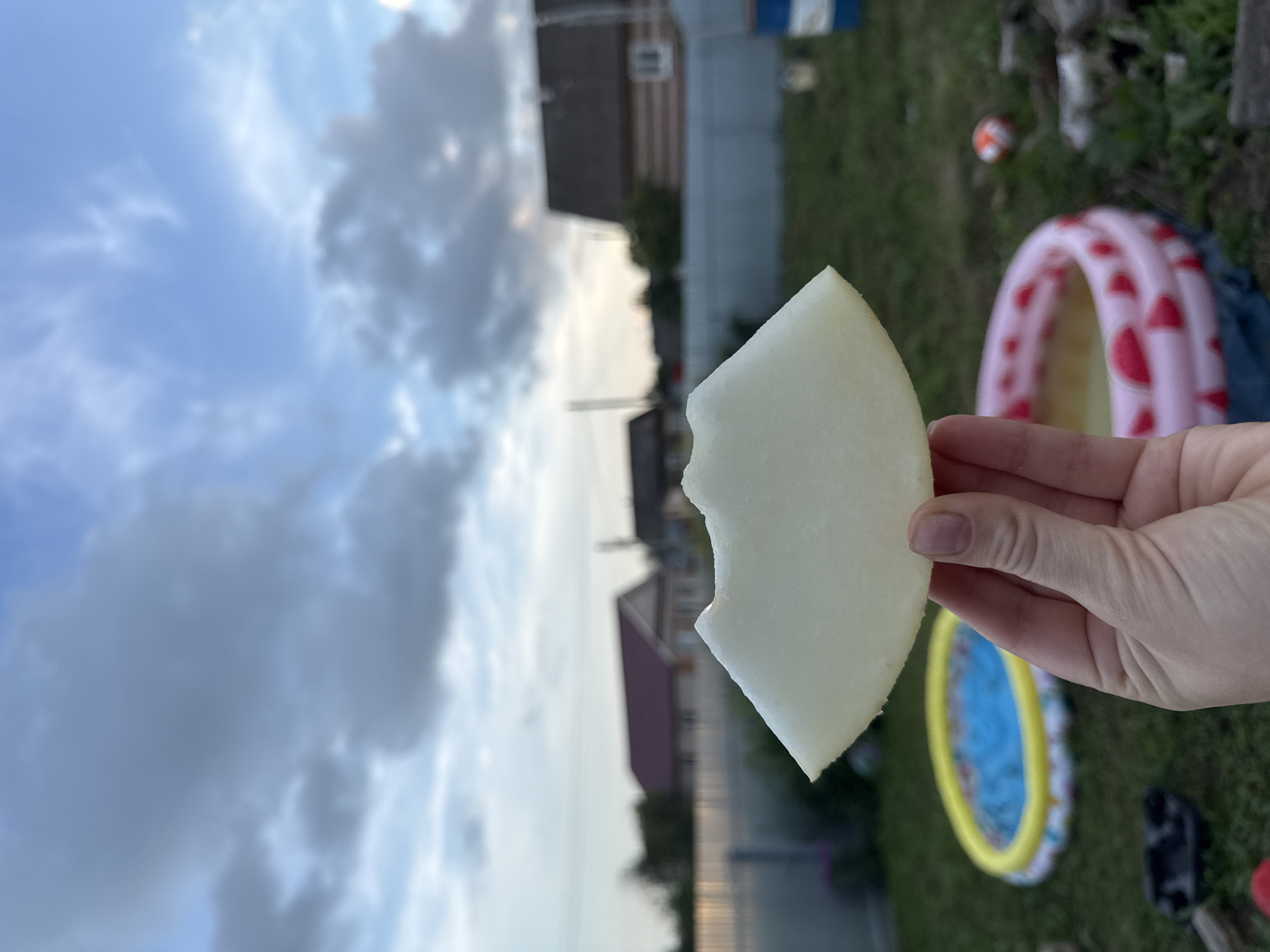 A hand is holding a slice of pale yellow melon with a bite mark out of it. The melon slice is in the foreground, with a blurred background of a backyard in Большая Уча, Russia. In the background, there are two inflatable kiddie pools, one blue and yellow, and another pink with red watermelon slices. There are also houses with red roofs and a grey fence visible. The sky is cloudy with some patches of blue.