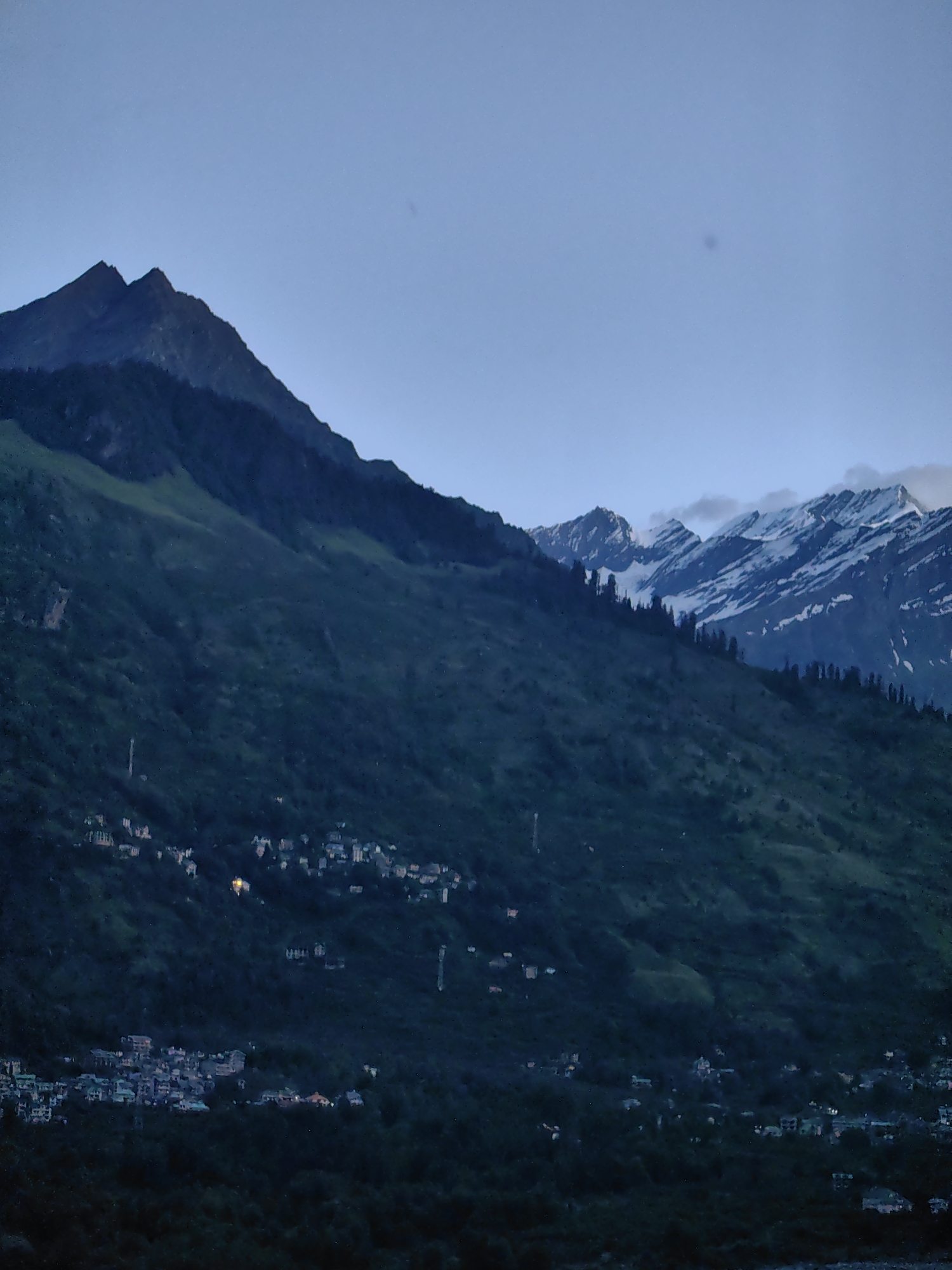The image shows a mountainous landscape at dusk, likely in or near Vashisht, India. The foreground is dominated by dark, densely forested hills, with a small village nestled within them. The village features numerous buildings with illuminated windows, suggesting human habitation. Above the forested hills, a larger mountain range is visible, some peaks of which are capped with snow. The sky is a pale, clear blue, transitioning into twilight. There are no visible people in the image. The overall impression is one of a serene, natural setting with a human settlement integrated into it.