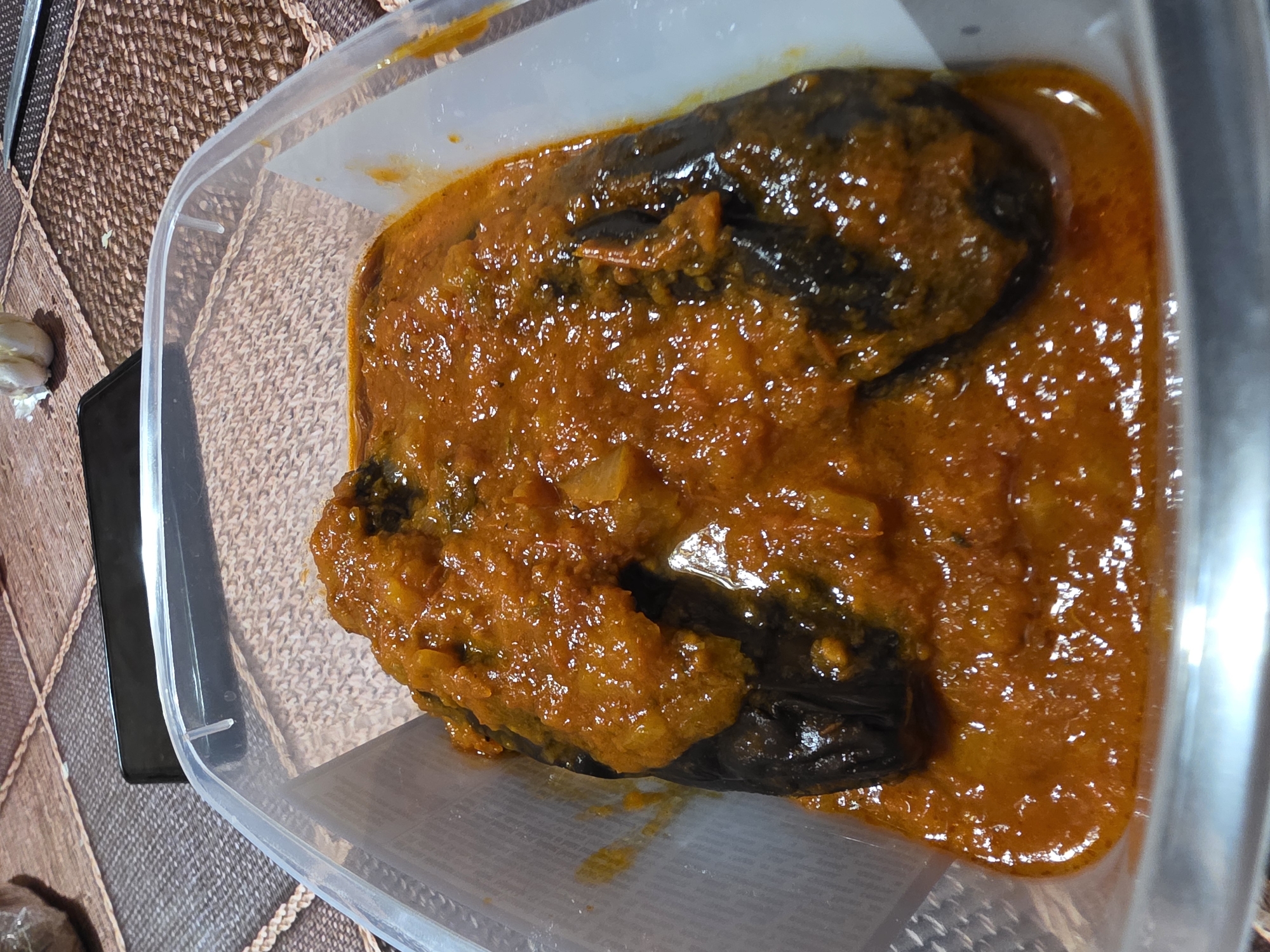 A close-up shot depicts a clear plastic container filled with food. The food appears to consist of pieces of a dark-colored vegetable, possibly eggplant, smothered in a thick, bright orange sauce. The surface of the sauce has a glistening, textured appearance. The container sits on a surface with a textured brown and beige patterned covering. A garlic bulb is visible in the upper left corner of the image. The location and details about people, actions, or interactions cannot be determined from the image. The city of Yagodovo, Bulgaria, is not visually confirmed in the image.