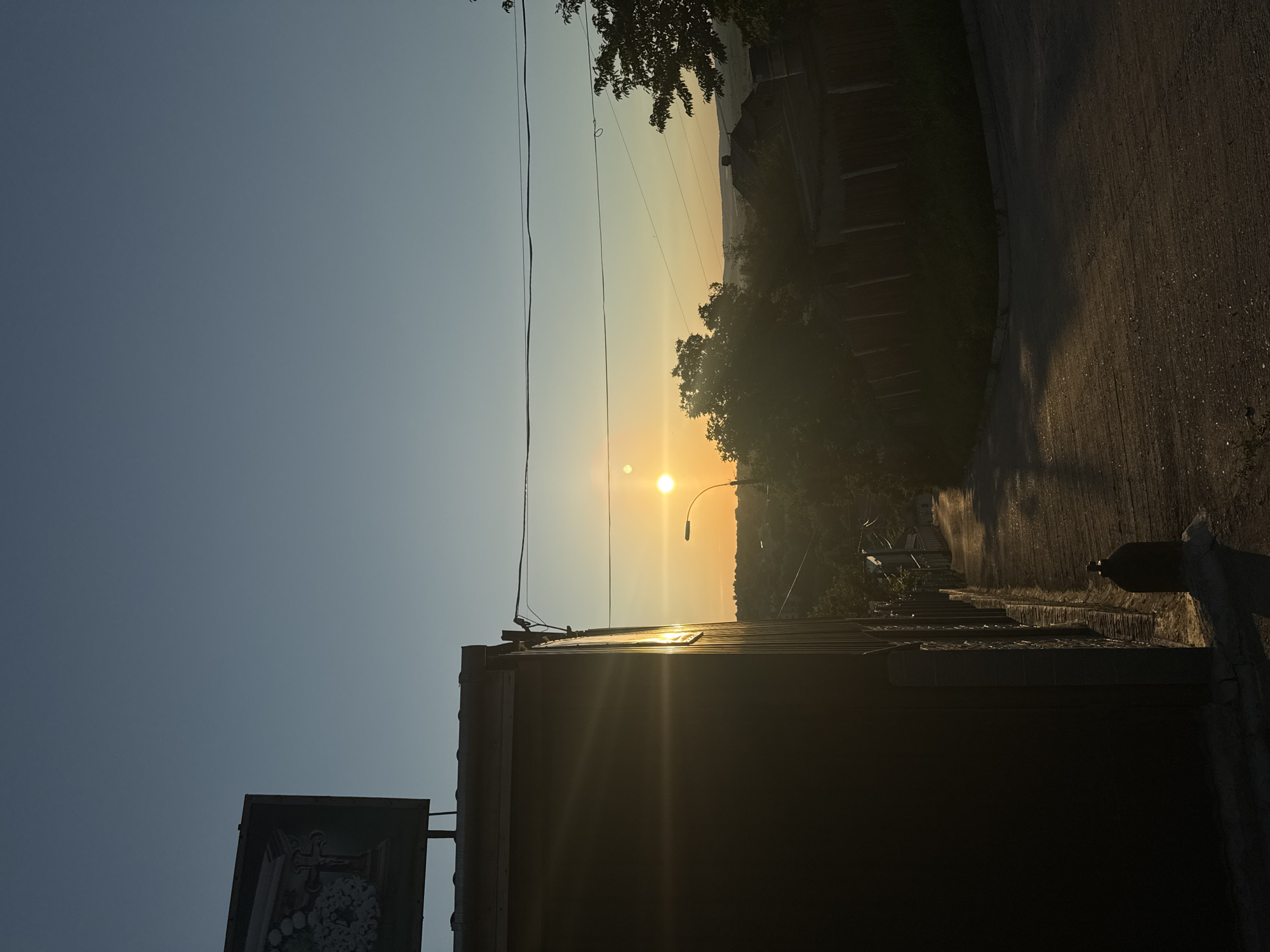 The image depicts a street scene in Strășeni, Moldova, during either sunrise or sunset, given the low angle of the sun and the warm, golden light casting long shadows. In the foreground, the left side of the frame is dominated by the dark side of a building, possibly a commercial structure, with a metallic roof and corrugated siding. A billboard with an image of a coffin adorned with flowers and a cross is affixed to the top left corner of this building.
The street itself is paved and slopes downwards and to the right. It is illuminated by the sunlight, creating streaks of light and shadow on its surface. To the right of the street, a low brick wall topped with wooden fencing lines the property of houses. Trees and vegetation are visible throughout the scene, suggesting a semi-urban or suburban environment.
In the middle ground, a lamppost stands on the right side of the street, its light diffused by the bright sun. The sun itself is a prominent feature, appearing as a bright, luminous orb in the center of the sky, with a smaller, fainter circle near it, potentially a lens flare or atmospheric effect. The sky transitions from a pale yellow near the horizon to a clear, pale blue higher up. The background shows rolling hills and the roofs of houses nestled amongst trees. Power lines are strung across the upper portion of the image, indicating the presence of infrastructure. There are no people visible in the frame. The overall impression is one of a quiet, residential area bathed in the serene glow of the sun.