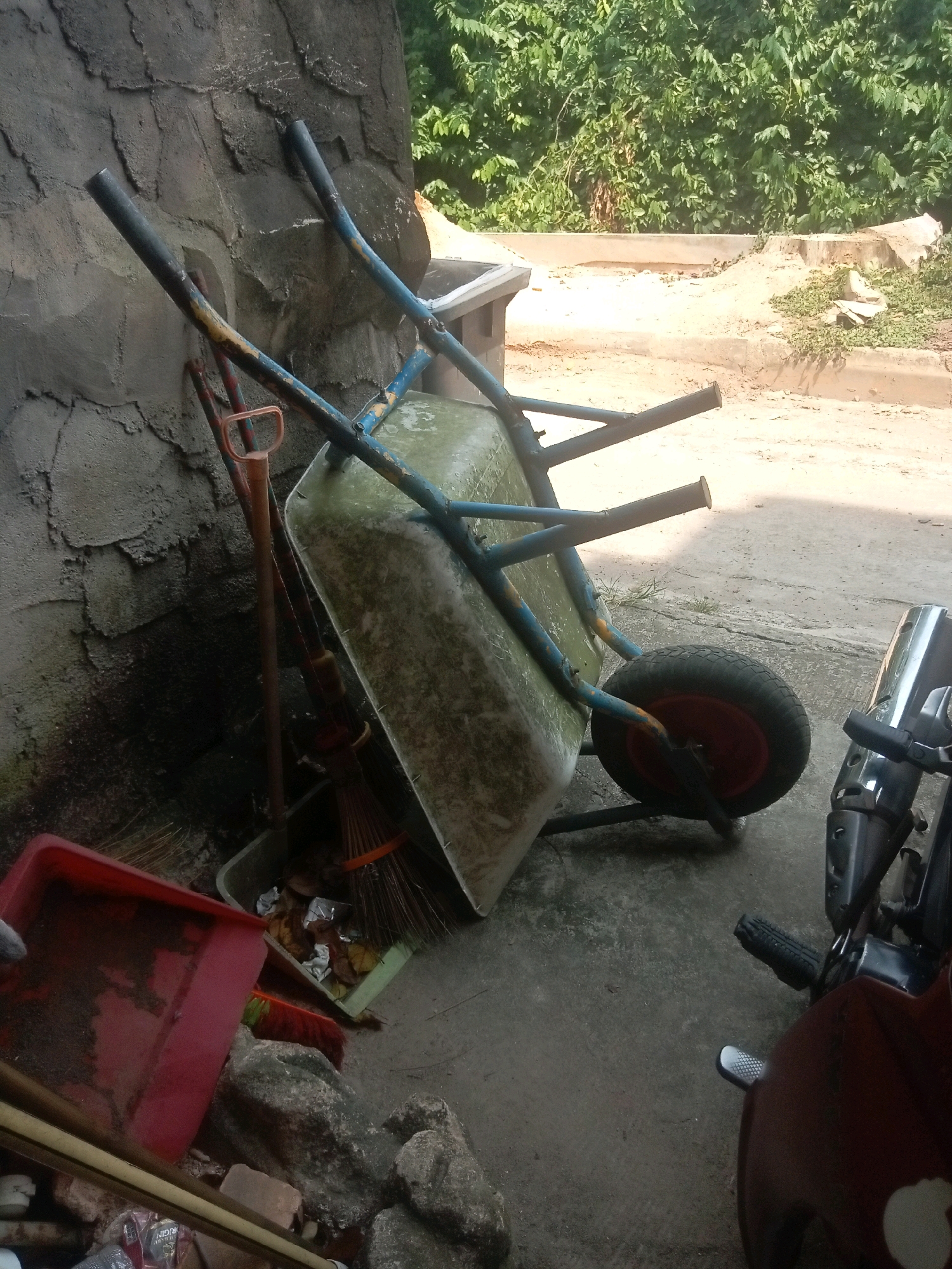 A wheelbarrow, a broom, a shovel, and a red plastic container are stacked against a textured, grey concrete wall. The wheelbarrow is tipped on its side, with its wheel facing right. A motorcycle is partially visible on the right side of the frame. The background features lush green foliage and a sandy, concrete-paved area. The scene appears to be an outdoor storage area in Batam, Indonesia, possibly a yard or workshop, given the presence of gardening and maintenance tools alongside a vehicle. The lighting suggests daytime.