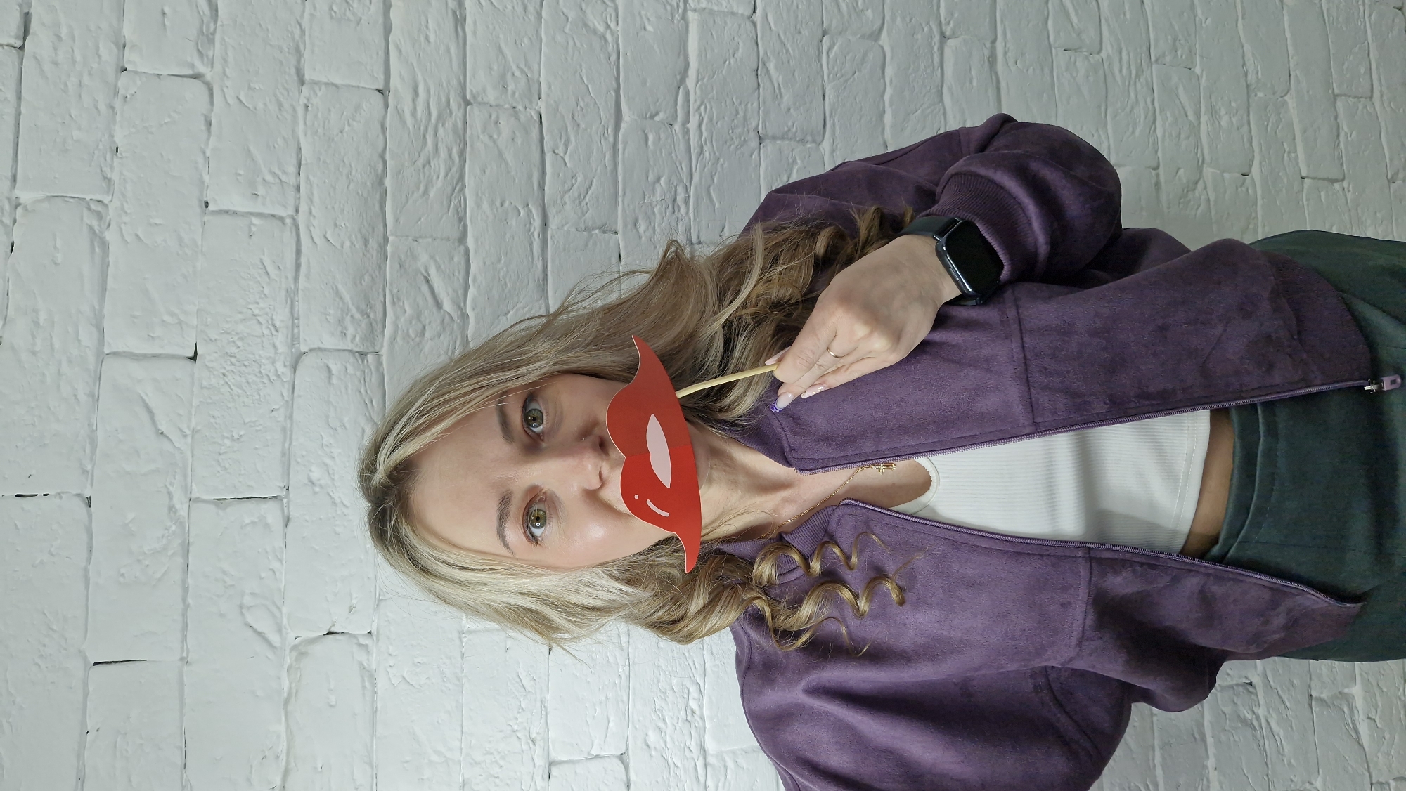 A blond woman with green eyes is holding a red cardboard cutout of lips on a stick near her face. She's wearing a purple zip-up jacket over a white top, with dark green pants visible. She has a black smartwatch on her left wrist and a gold necklace. The background is a white brick wall. The image is oriented vertically. No location is evident from the image.