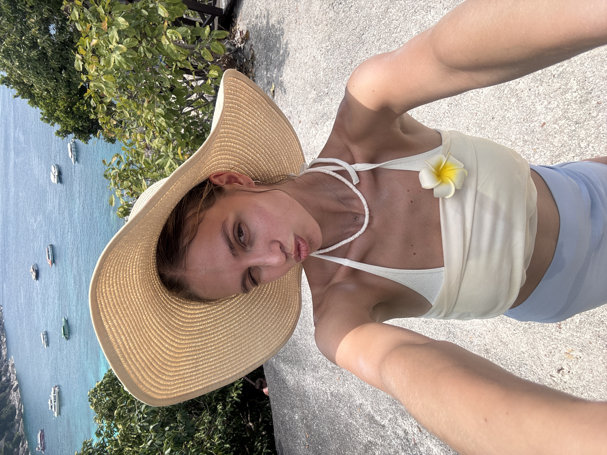 A young adult female takes a selfie outdoors in Lam Kaen, Thailand. She wears a large straw hat, a white top with a frangipani flower, and blue shorts. The background includes a body of water with boats, greenery, and a concrete surface.