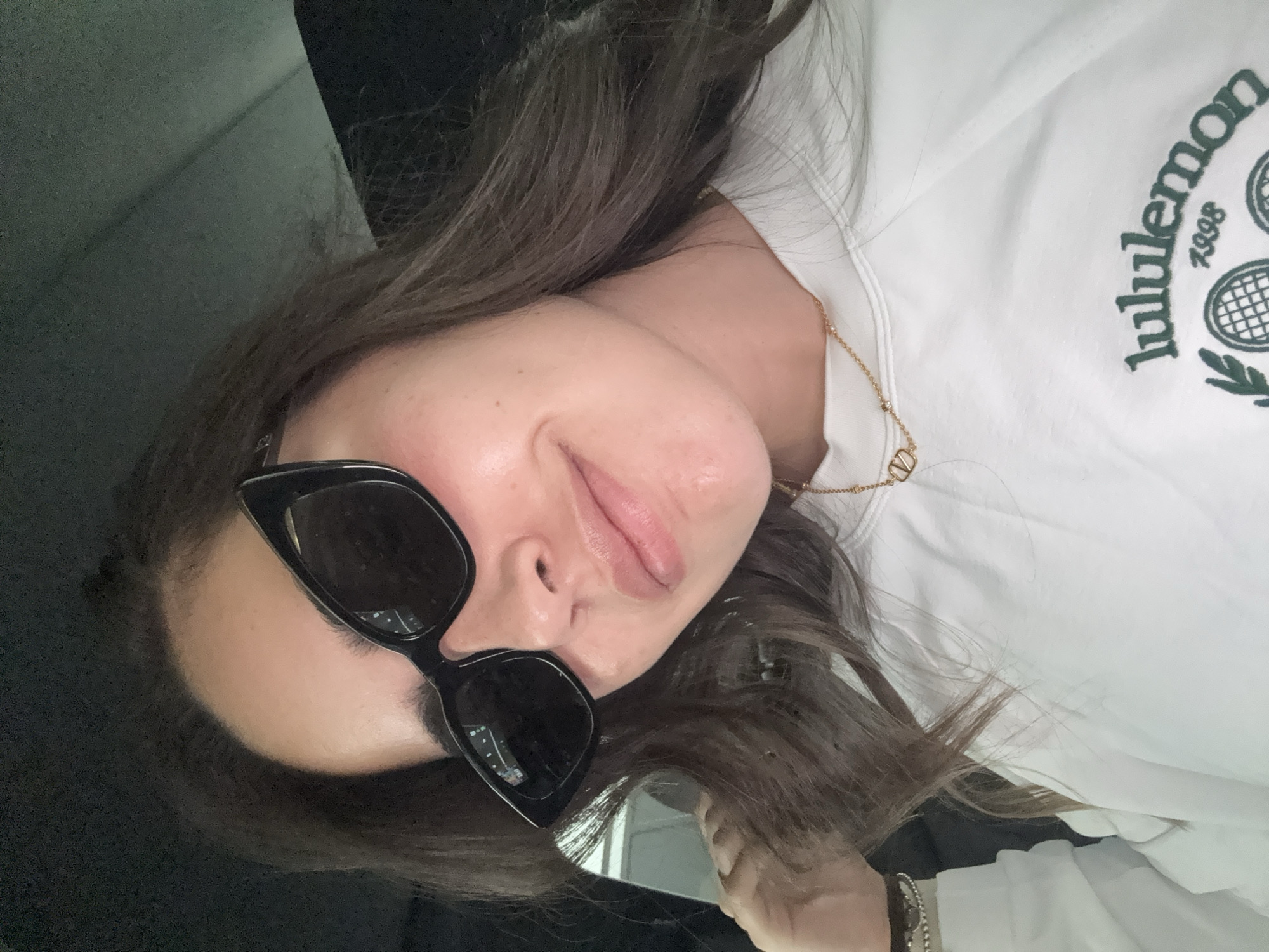 A woman with brown hair is wearing sunglasses and a white Lululemon t-shirt with a gold necklace. The location of the photograph is not discernible from the image.