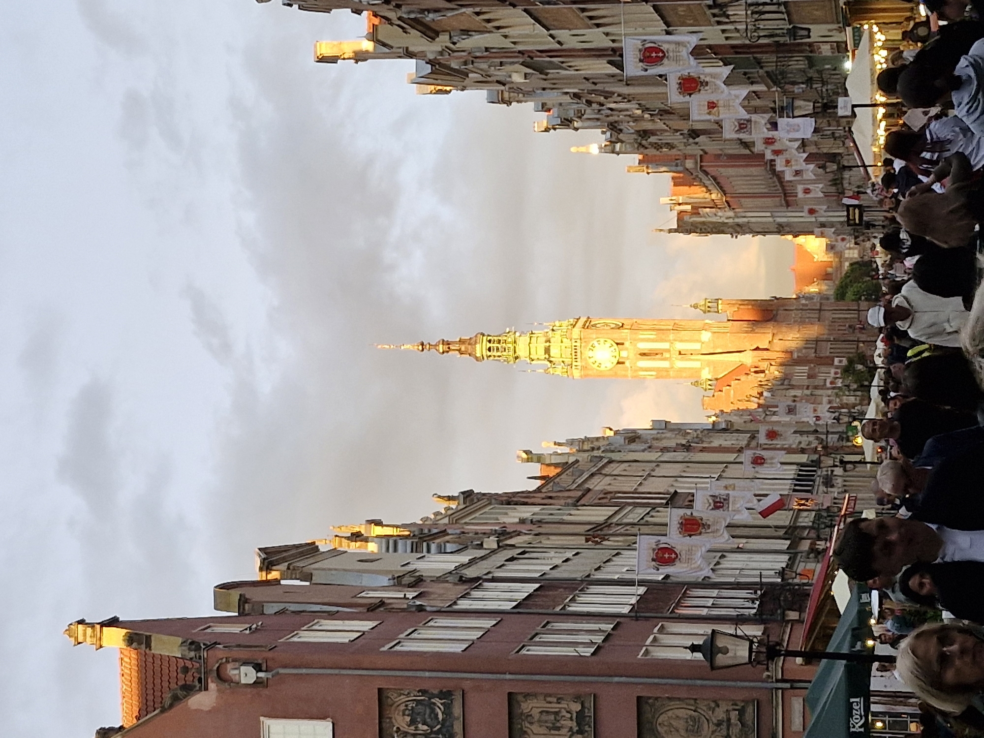 This is a vertical, eye-level shot of a bustling street scene in Gdańsk, Poland, likely Długa Street, leading towards the Main Town Hall. The image captures the golden hour, with warm sunlight illuminating the ornate facades of the historic buildings and casting long shadows.
The architecture is characteristic of Gdańsk, with tall, narrow buildings adorned with intricate gables and colorful plasterwork. Many of these buildings are decorated with hanging banners, some featuring coats of arms.
Dominating the background is the impressive tower of the Main Town Hall, its clock face clearly visible and illuminated by the setting sun. The sky above is partly cloudy, with a mix of grey and soft, illuminated clouds.
The street is filled with a diverse crowd of people, suggesting a lively atmosphere, possibly a festival or a busy evening. Individuals are walking, talking, and looking around, creating a sense of movement and activity. Some are clustered around food stalls or shops visible along the street.
In the foreground, several people are in focus, their faces and attire visible, while others are more blurred, indicating their movement or distance from the camera. Among the notable foreground elements is a lamppost with an old-fashioned lantern on the left side of the image, and green awnings of outdoor seating areas.
