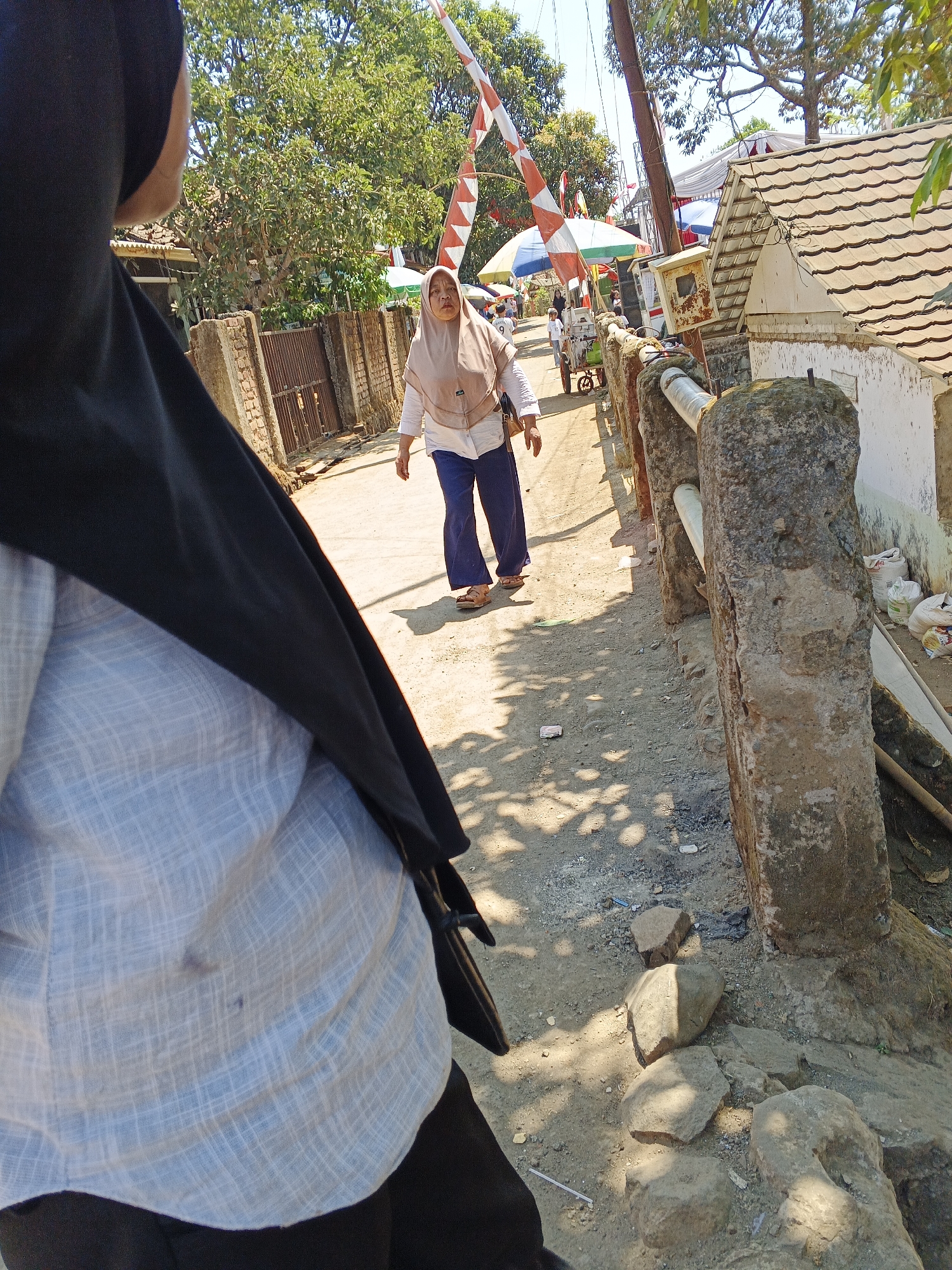 The image captures a street scene in Sukaharja, Indonesia. In the foreground, a person wearing a light blue patterned shirt and a black hijab is partially visible, their back to the camera. Further down the dirt road, a woman in a light brown hijab and white shirt, with dark blue pants and brown sandals, walks away from the camera. She is looking to her left with an open mouth. Behind her, a red and white striped barrier is raised, and a small cart with an umbrella is visible. The environment is rustic, with brick walls, trees, a building with a tiled roof, and a stone railing along the right side of the road. The ground is dusty, with scattered rocks and debris. The sun is shining brightly, casting shadows.