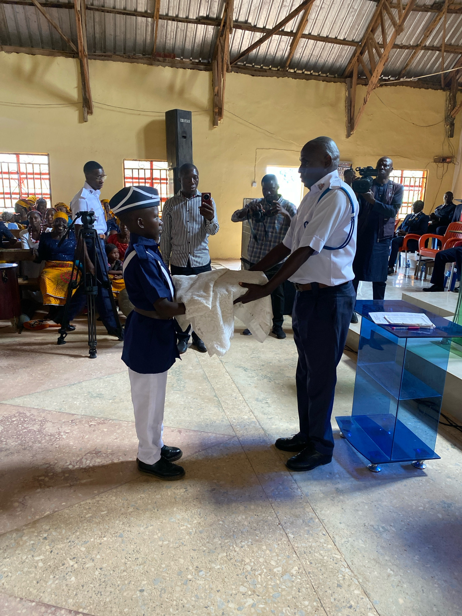 In a hall in Binchi, Nigeria, a young boy in a navy blue uniform with a white shirt and a peaked cap stands holding a white cloth. He is facing right, and his feet are clad in black shoes. To his right, an adult man in a white shirt with blue trim, a navy blue sash, and dark trousers stands with his hands on the cloth. This man is also wearing black shoes. Behind them, a camera operator with a video camera is visible, as is a man holding a red mobile phone. In the background, an audience sits on wooden benches, some women wearing brightly colored headscarves. The hall has a corrugated metal roof supported by wooden beams, and yellow walls. A blue acrylic podium stands to the right of the man in the white shirt.