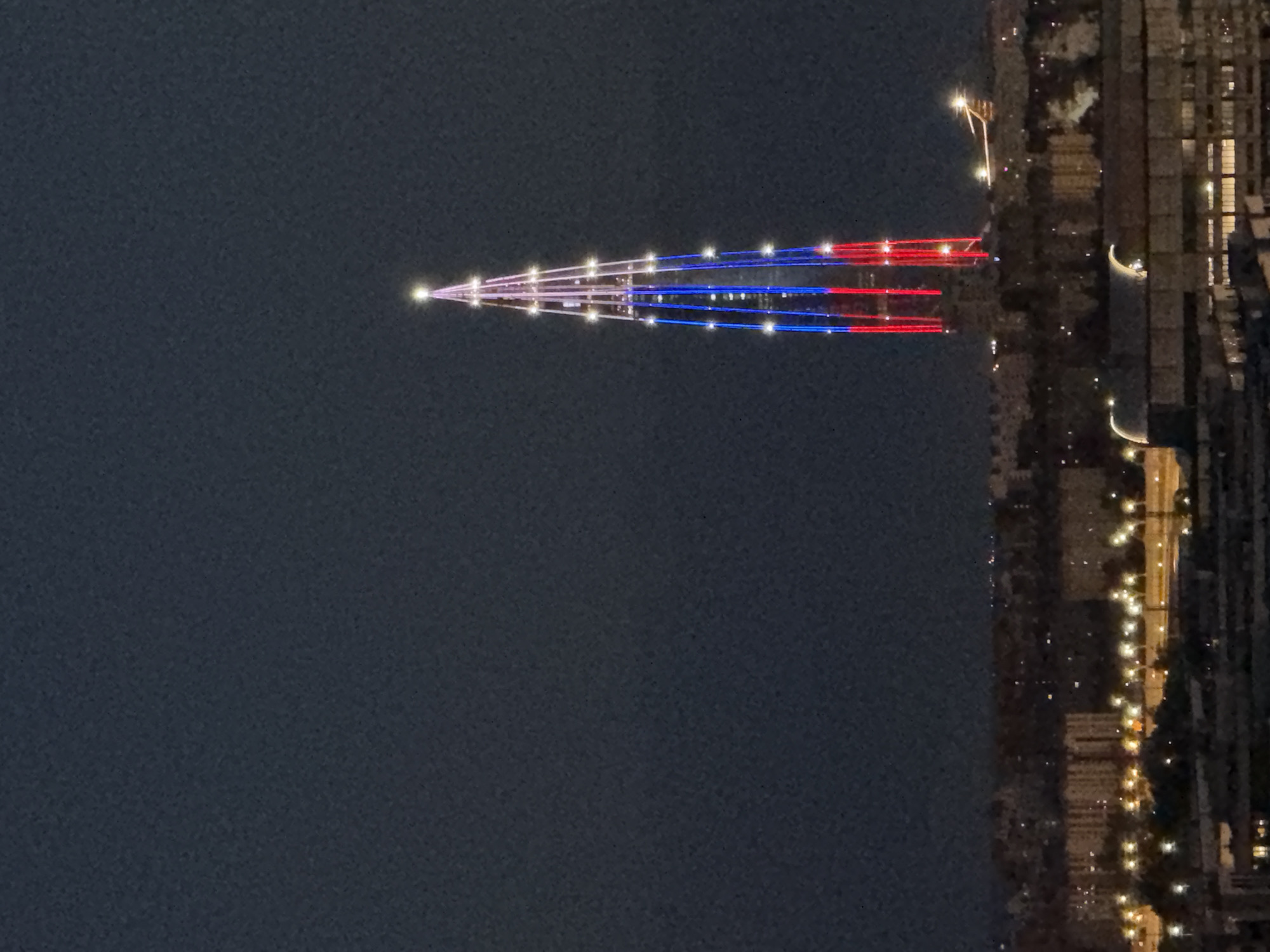 This image captures the Lakhta Center skyscraper in St. Petersburg, Russia, illuminated at night. The skyscraper is lit with the colors of the Russian flag: red, white, and blue. The top of the building is adorned with a bright white light. The background consists of the dark night sky and a silhouette of the city below, with scattered lights from buildings and streetlamps. The image is taken from a distance, showing the vastness of the urban landscape and the prominent, brightly lit structure. There are no people or specific actions depicted in the image; it is a static nighttime view of the city and its landmark skyscraper.