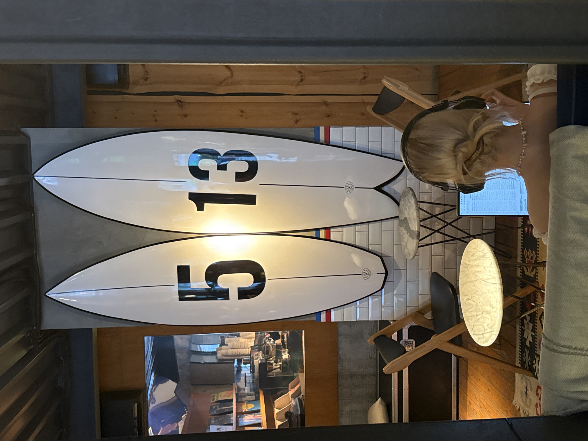 This image, set indoors in Moscow, Russia, features a person and decorative surfboards within a modern, surf-themed interior. The scene is lit by artificial light, suggesting evening or nighttime.
Two prominent white surfboards with black outlines are mounted vertically on a wall of white subway tiles. The upper surfboard clearly displays the large black number "13," while the lower one features the number "5." A thin black line runs down the center of each board, and a small, circular emblem is visible near the tail of the lower surfboard.
To the right, a person with blonde hair is seated, facing away from the viewer, wearing black over-ear headphones. They are focused on an illuminated screen, likely a laptop or tablet, placed on a small, round table with a light, marble-patterned top. This suggests the person is engaged in work, study, or entertainment.
The furniture includes minimalist wooden chairs with black seats. The surrounding decor incorporates light wooden planks, a dark metallic structure, and a patterned rug with geometric designs. A reflection in a nearby mirror or glass surface reveals a bookshelf and other interior details. The overall ambiance is cozy and contemporary, hinting at a cafe, co-working space, or a leisure establishment with a unique design inspired by surf culture.