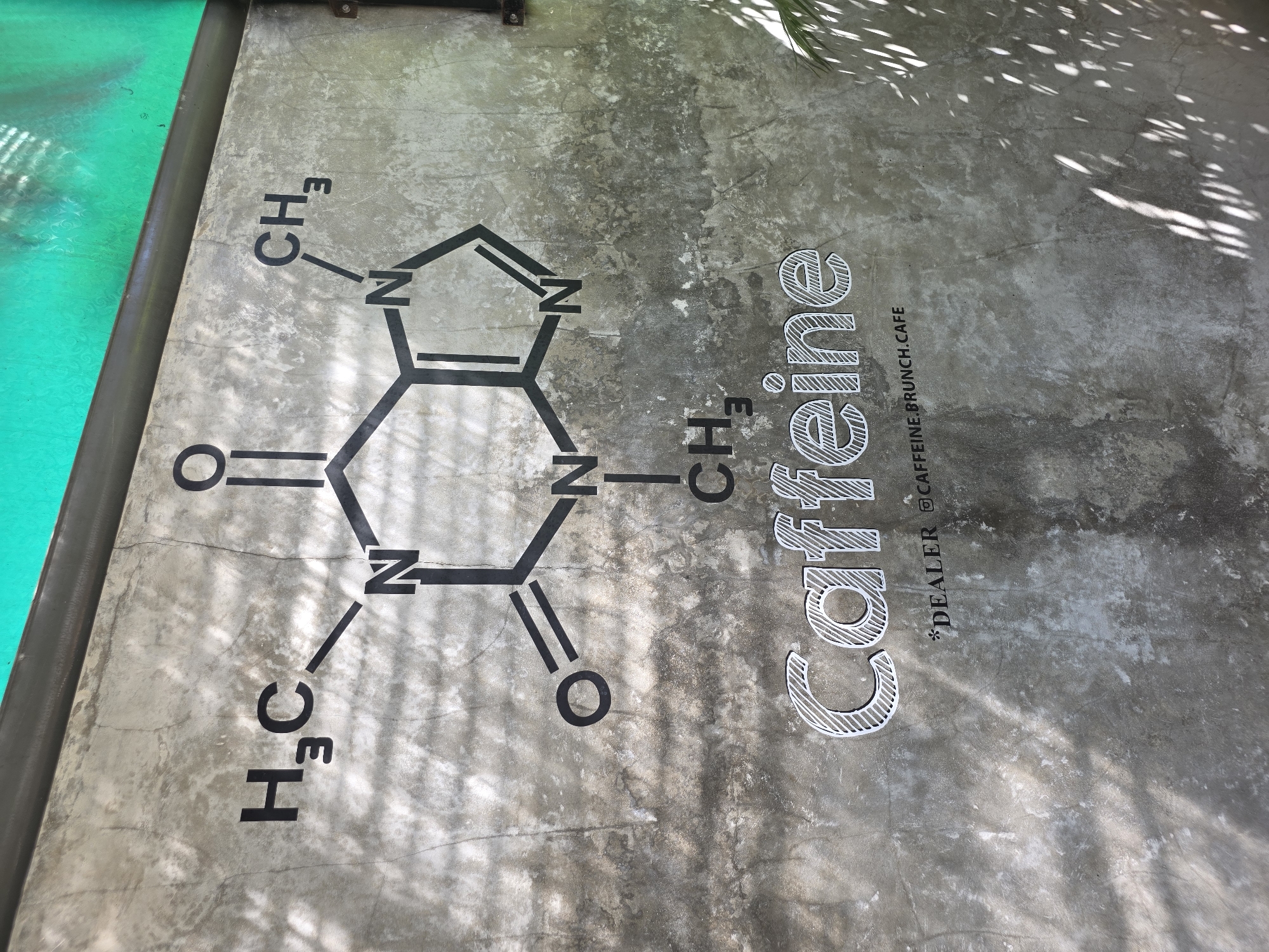 The image shows a concrete surface with a depiction of the caffeine molecule and the word "Caffeine" in a stylized, lined font. Below the word "Caffeine" is the text "*DEALER @CAFFEINE.BRUNCH.CAFE". The background texture suggests a cafe setting. Based on the text, the location is likely Caffeine Brunch Cafe in North Kuta, Indonesia.