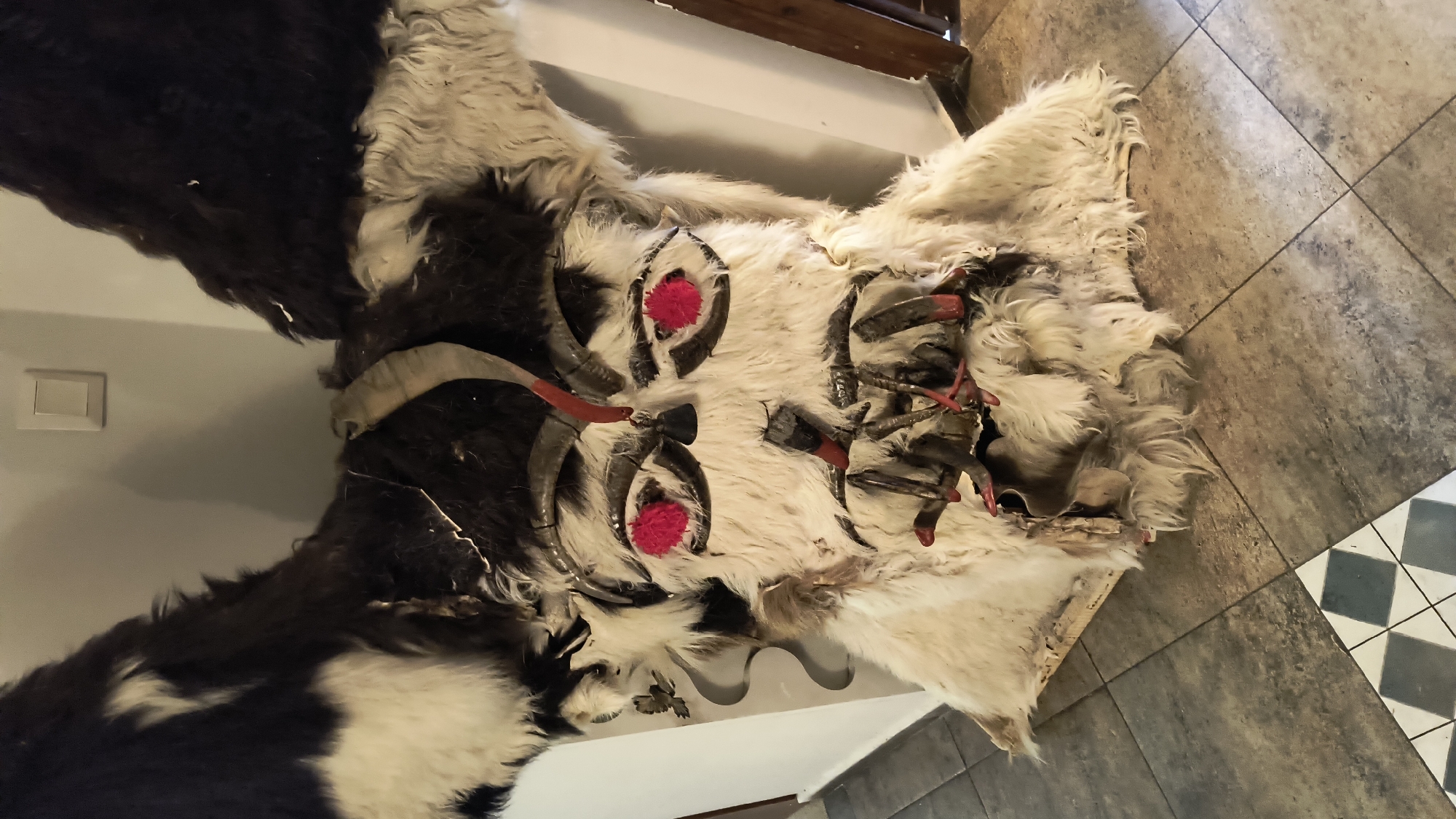 The image depicts a traditional Bulgarian Kukeri mask. The mask is constructed from animal hide, primarily white fur with patches of black. It features decorative elements such as horns, possibly from a goat or ram, small axes, and red pom-poms in place of eyes. The mask is resting on a surface that includes a wall, visible light switch, and tiled floor. The specific location within Sofia, Bulgaria, is not discernible from the image alone.