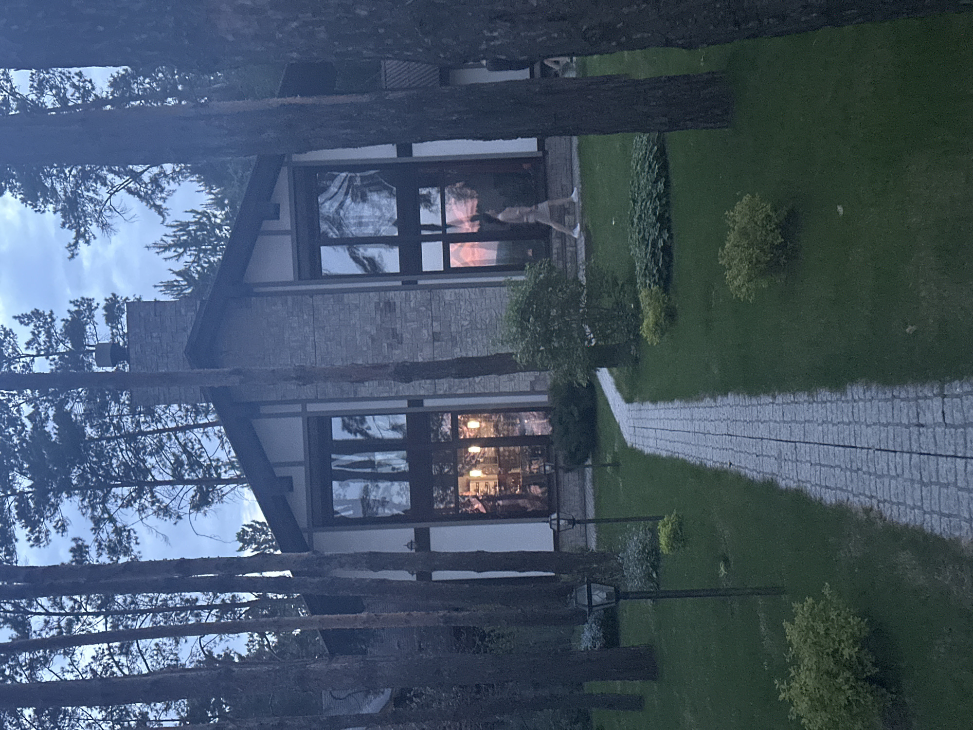 A ground-level, wide-angle shot depicts the exterior of a two-story house, seemingly constructed of stone and featuring large, dark-framed windows. A stone pathway leads towards the house, bordered by a neatly manicured lawn with shrubbery. Several tall, thin trees are situated in the foreground, partially obstructing the view of the house. The time of day appears to be dusk or early evening. Reflected in one of the windows is a person in light-colored clothing standing. The house has a steeply pitched roof. Based on visual cues in the window reflections, it is inferred that the location is in Aviator, Russia.