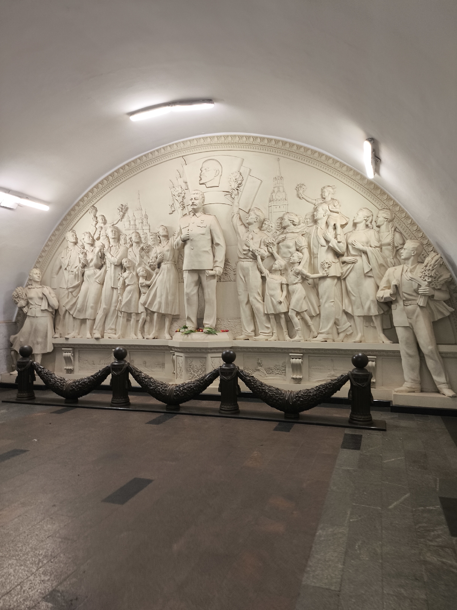 The image shows a large, white bas-relief sculpture depicting a group of people, including a prominent figure in a military uniform who appears to be Stalin, along with a likeness of Lenin in the background. The group is carrying flowers and appears to be celebrating. The sculpture is located in a subway station, likely in Russia, based on the style of the art and the subject matter. The setting is underground, suggested by the arched ceiling and artificial lighting. A dark metal decorative barrier is in front of the sculpture. The floor has mixed grey and brown tiles. There are a few flowers placed at the base of the sculpture of Stalin.