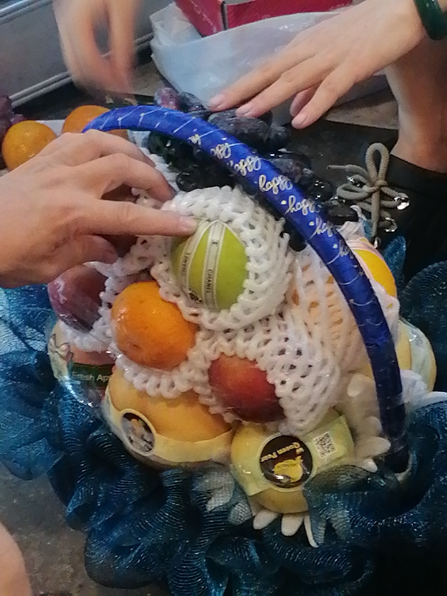 A close-up shot captures a fruit arrangement, possibly a gift basket, with several fruits like pears, oranges, and possibly apples nestled within white foam netting. A blue ribbon with white cursive text, possibly a celebratory message, arches over the fruits. The arrangement is placed on a dark, textured surface, possibly a table, with a dark blue mesh material underneath. In the background, blurred hands are visible, suggesting human interaction with the fruit arrangement, and parts of what appear to be a white plastic bag and a red object are also present. The overall scene suggests a commercial setting, possibly a market or a shop, in Ho Chi Minh City, Vietnam, where such gift arrangements are common.