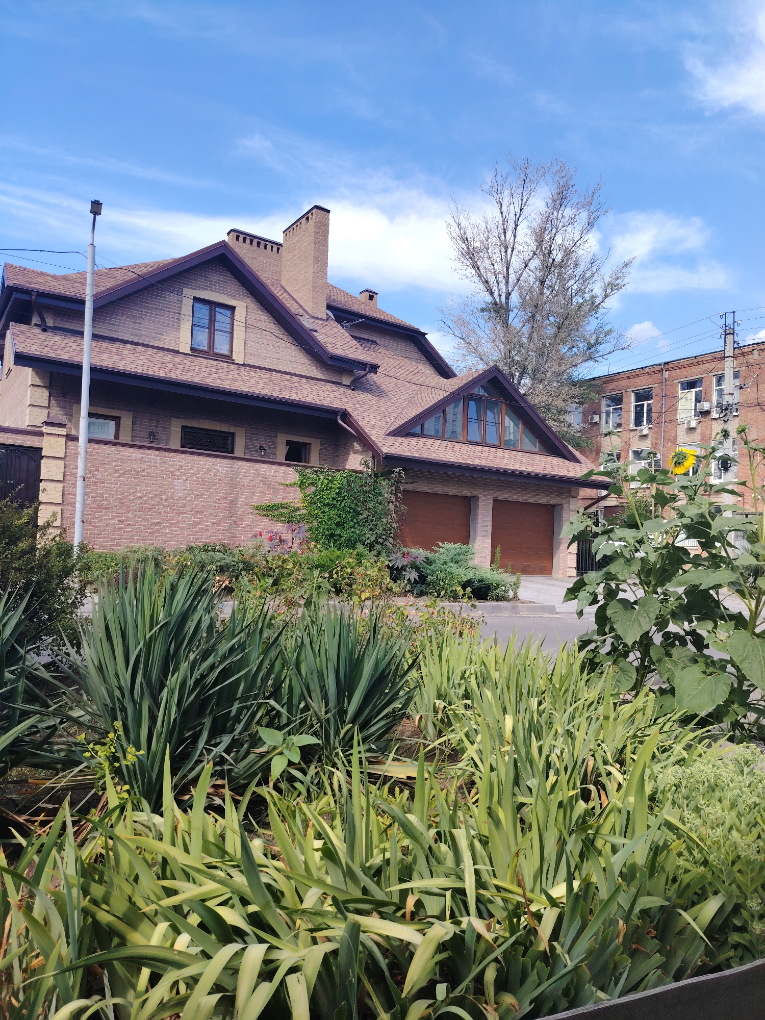 The image captures a residential property on a sunny day in Rostov-on-Don, Russia. A large, two-story house with a brown tiled roof and brick facade dominates the center of the frame. The house features a prominent brick chimney and several windows, including a large triangular window under the roof. In front of the house, there are two garage doors. The foreground is filled with lush green vegetation, including tall, spiky plants and a sunflower in bloom to the right. Behind the house, a multi-story brick building, possibly an apartment or office building, is visible. Power lines and a street lamp are also present. The sky is a clear blue with scattered white clouds. There are no people visible in the image.