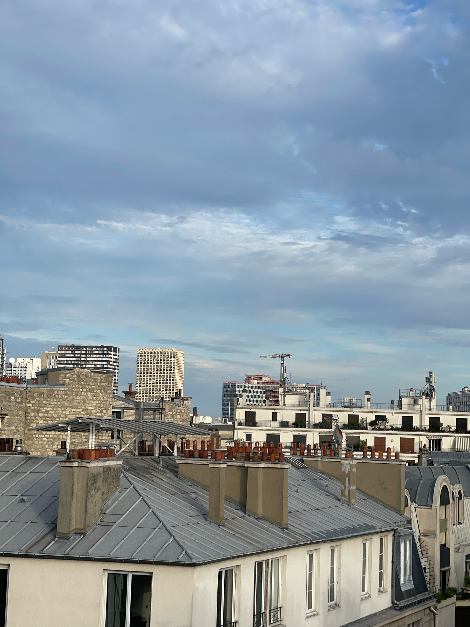 This is a medium-shot, eye-level, outdoor, daytime, vertical photograph of a cityscape in Paris, France. The foreground is dominated by the zinc-covered rooftops of Haussmannian-style buildings. These rooftops are characterized by their steeply pitched, grey metal surfaces, punctuated by numerous red-brick chimneys, some of which are capped with terracotta pots. Several chimneys are also visible, made of stone and plaster. The buildings themselves are rendered in a light color, with cream-colored facades and numerous white-framed windows. Some of the windows are open, revealing dark interior spaces.
Behind these foreground rooftops, the middle ground of the image reveals a more modern urban landscape. A large, light-colored building with a grid-like pattern of windows is prominent, as is another taller, darker building with horizontal bands of windows. A construction crane is visible in the distance, suggesting ongoing development. Other buildings in this section are white, with multiple stories and balconies, some adorned with greenery. Antennas and satellite dishes are visible on some of these rooftops.
The background of the image is a wide expanse of cloudy sky. The clouds are a mix of light grey and white, with some darker patches, indicating an overcast or partly cloudy day. The lighting suggests it might be early morning or late afternoon, as the light is diffused and soft. There are no people visible in the photograph. The scene depicts a typical Parisian urban environment, showcasing a blend of historic architecture and modern construction.