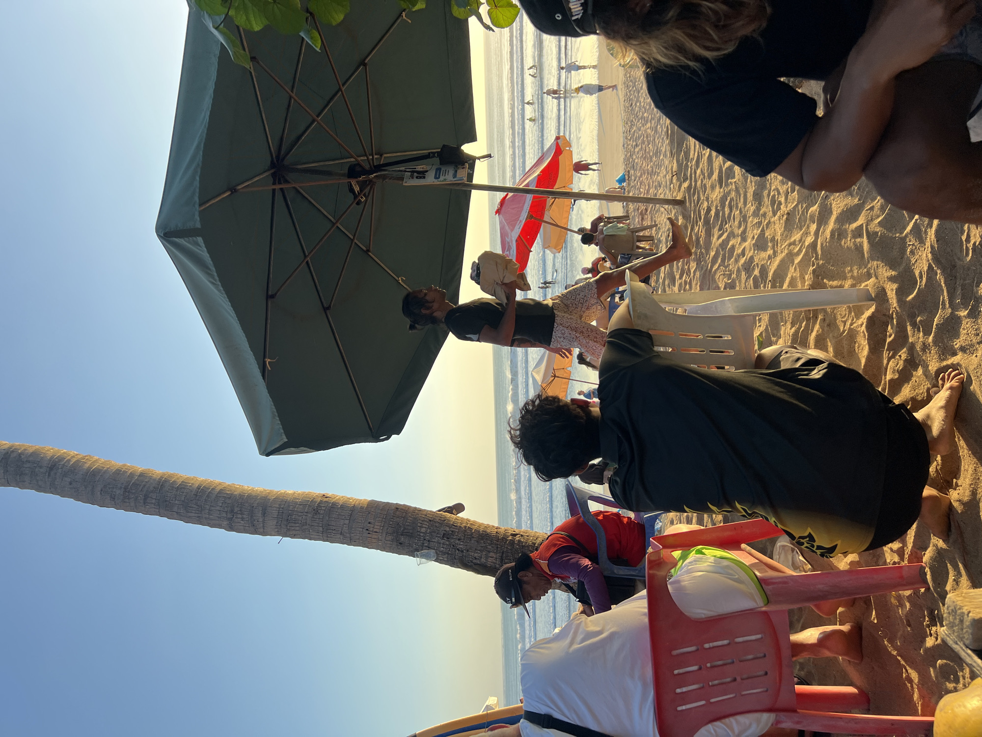 The image depicts a beach scene in Kuta, Indonesia, during what appears to be late afternoon or early evening, with the sun casting long shadows. In the foreground, several people are seated on the sand at tables, partially obscured by a large green umbrella. One person is seated on a red plastic chair, facing away from the camera. Another person, also facing away, is wearing a dark t-shirt with yellow accents and black shorts. To the right, a person in a black t-shirt and shorts is partially visible, with long blonde hair.
A man walks in the middle ground, carrying a bundle wrapped in brown paper. He is wearing a dark t-shirt and patterned shorts. Behind him, people are scattered on the beach, some wading or swimming in the ocean, which has moderate waves. There are several beach umbrellas, including a red and white one. A tall palm tree trunk is prominent on the left side of the image, leaning slightly towards the right. The ground is covered in golden sand. The sky is a clear, pale blue.