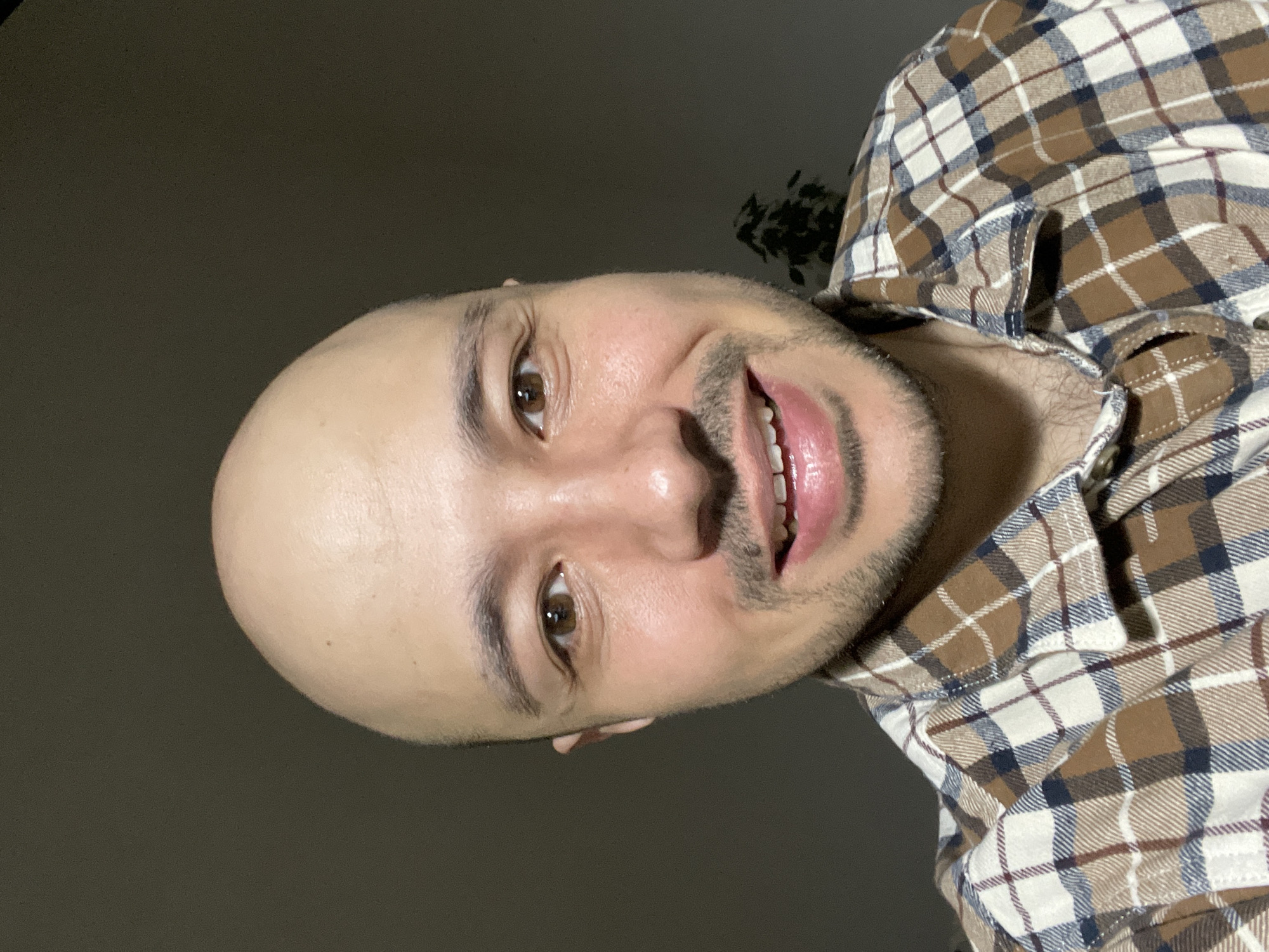 A bald man with brown eyes and a short beard is smiling at the camera. He is wearing a tan, brown, white, and blue plaid shirt. The background is a dark gray wall. There is also a plant in the background behind the right shoulder of the man. There is no evidence to suggest that the location is Izhevsk, Russia.