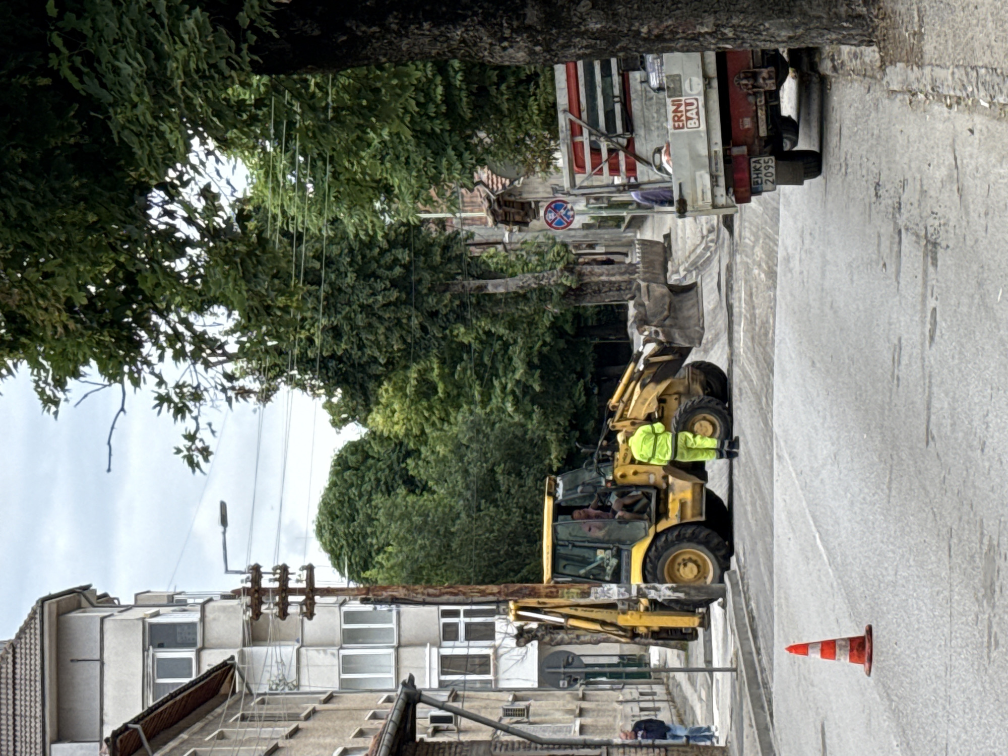 The image depicts a street scene in Pleven, Bulgaria, likely undergoing road construction. In the foreground, a bright orange traffic cone with white stripes is positioned on the asphalt. To the left, a yellow backhoe loader is parked, with two individuals visible inside the cabin. A man in a high-visibility yellow jacket and pants stands next to the backhoe, facing away from the camera. Further down the street, a red truck with a flatbed is parked on the right side of the road. The truck has the text "ERNI BAU" and a license plate with the inscription "EHKA 2095". Behind the truck, a blue and white circular sign is visible on a pole. Buildings line both sides of the street, with trees providing a green canopy overhead. The sky is overcast.