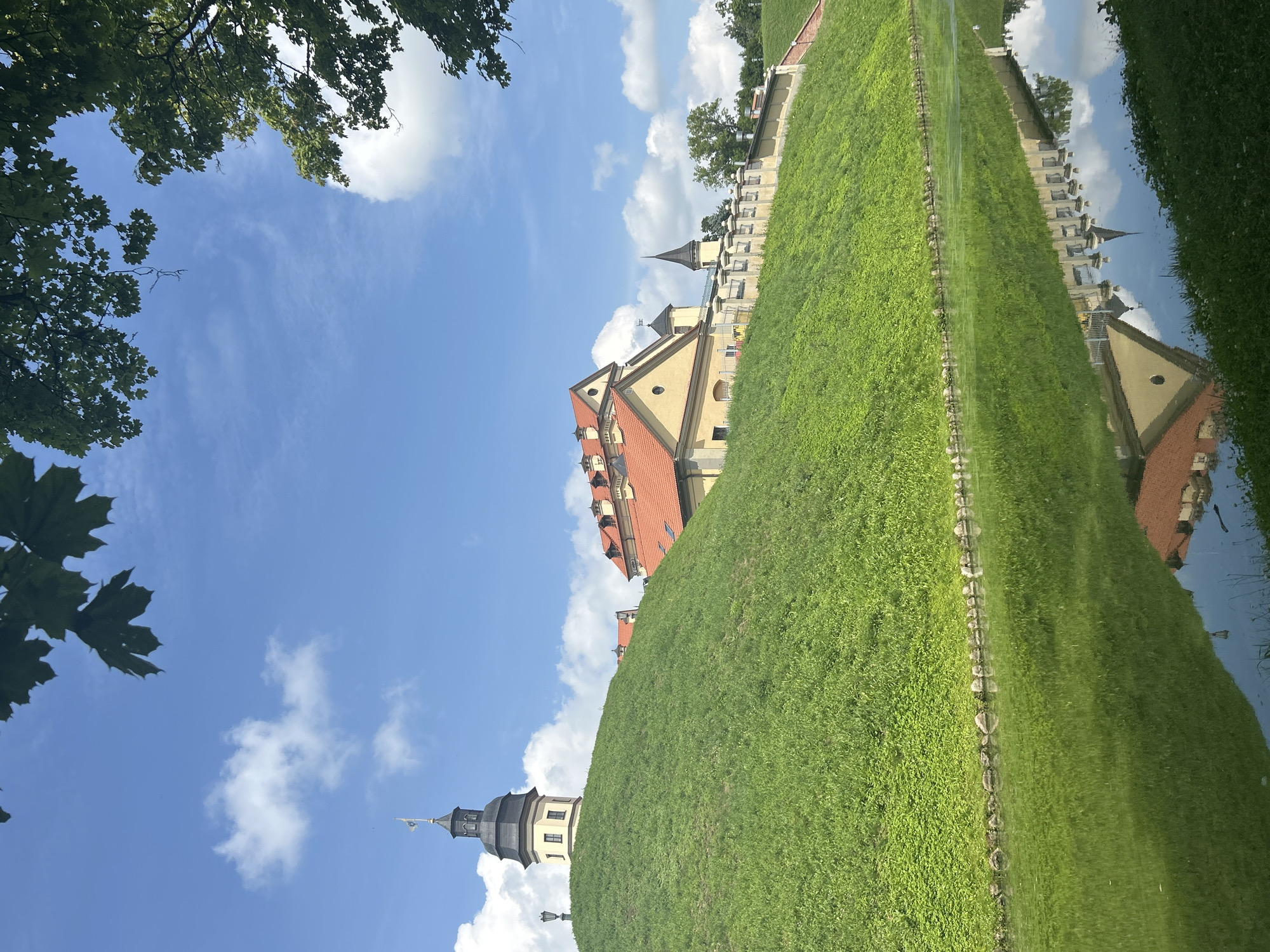 The image shows the Nesvizh Castle complex in Nesvizh, Belarus, on a sunny day with a blue sky and scattered clouds. In the foreground, a calm body of water reflects the castle and the surrounding grassy hill. A steep, verdant hill dominates the middle ground, leading up to the castle buildings. The castle itself is a large, yellow structure with red tiled roofs and several towers, including a prominent one with a black cap and a flag. A low, white stone wall with decorative elements runs along the top of the hill, separating the castle from the surrounding landscape. Lush green grass covers the hill and the bank of the water. In the upper right corner, the dark green leaves of a tree frame the scene. The reflection in the water is clear, showing a mirror image of the castle, the hill, and the sky.