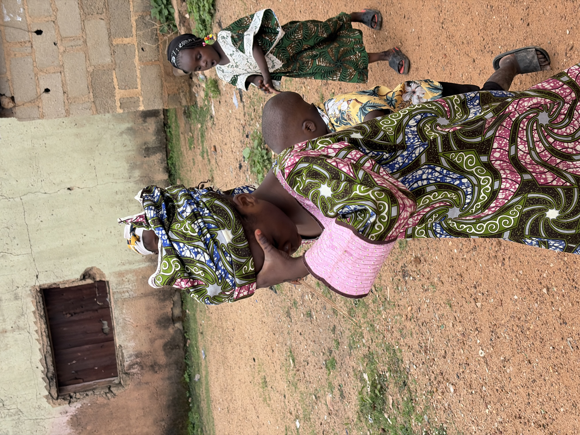 The scene is set in an outdoor area, possibly a yard, in the city of Barakin Lerawi, Nigeria. The ground is covered in reddish-brown dirt with sparse patches of green vegetation. In the foreground, a woman is breastfeeding a child. Both the woman and the child are wearing traditional patterned African clothing. Another child, wearing a green patterned dress with white lace, stands in the background. Adjacent to the group is a building with a weathered, light-colored wall. A small, boarded-up window is visible on the wall. The scene is captured in natural daylight.