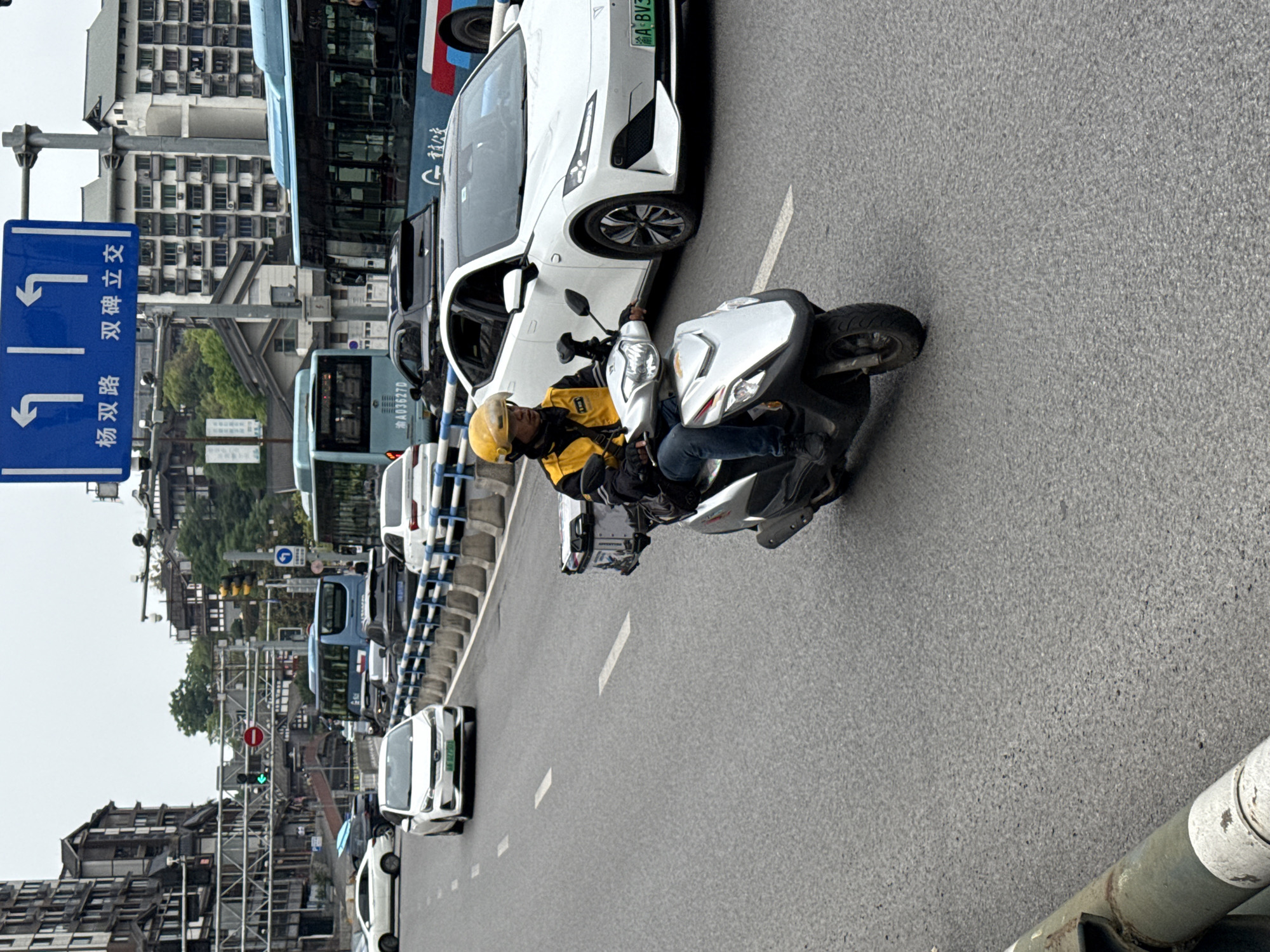 The image shows a street scene in Shapingba District, China. A delivery driver wearing a yellow helmet is on a white scooter in the middle of a busy road with several cars and buses around. The road has painted white lane markings. A blue road sign displaying Chinese characters is visible near the left side of the image. Buildings are visible in the background. A traffic light shows a green light.