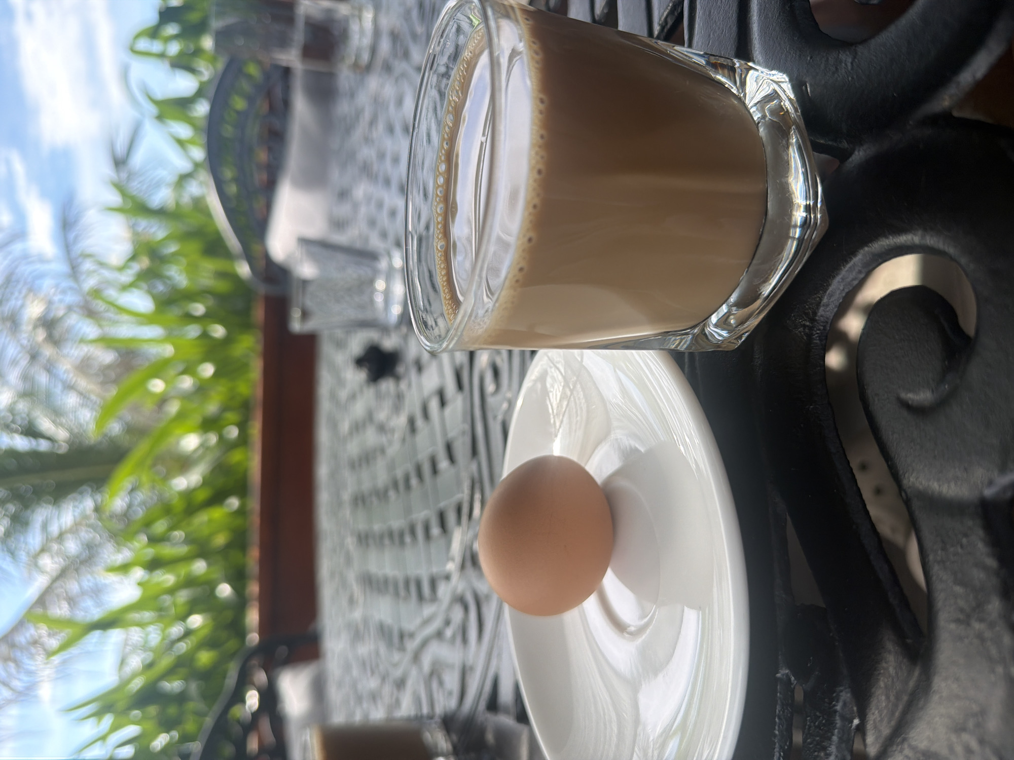 An overhead shot shows a breakfast setting on a black metal table. In the foreground, a single brown egg rests on a white saucer. To its right, a clear glass holds a brown, frothy beverage. The background is blurred, showing lush green foliage and a bright sky, suggesting an outdoor dining area in Bắc Nha Trang, Vietnam.