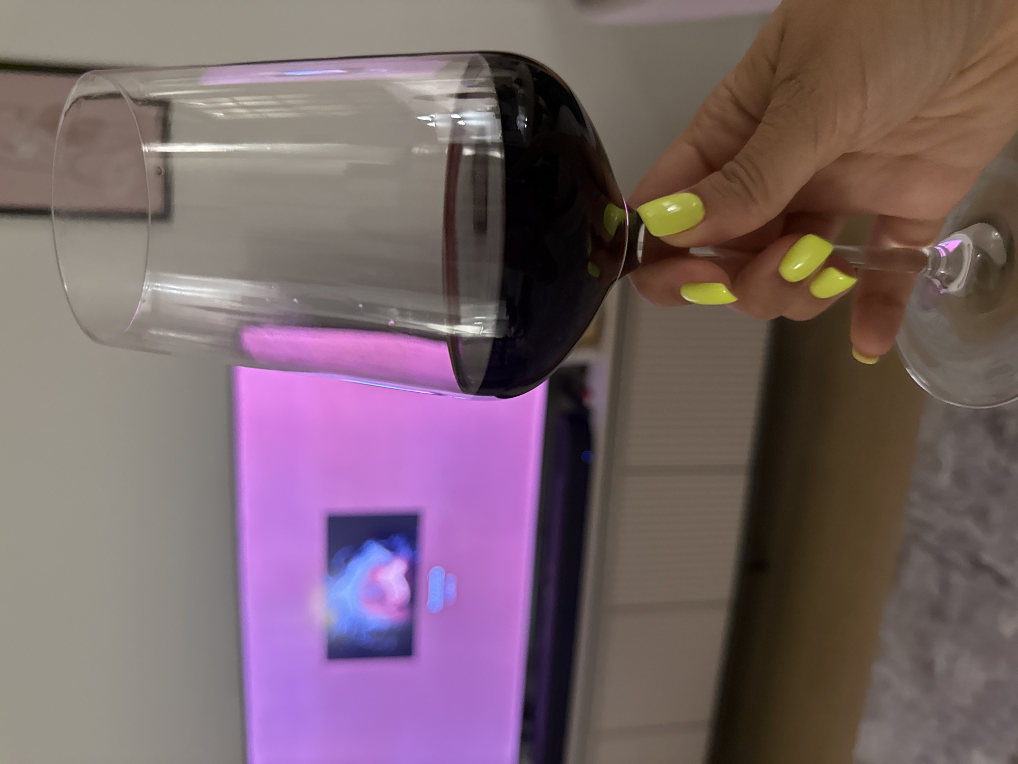A hand with yellow nail polish is holding a wine glass filled with red wine. A television screen showing an image of a nebula is visible in the background. The location and city are unknown.