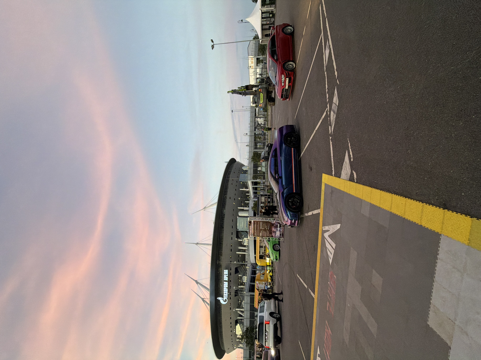 In a parking lot facing the Gazprom Arena in St. Petersburg, Russia, a serene dusk scene unfolds under a pale pink and blue sky. Several cars are parked, including a prominent iridescent purple Nissan GT-R, a red Mitsubishi Lancer Evolution, and a white SUV. In the background, a yellow food truck and a green car add splashes of color. The Gazprom Arena, with its distinctive sail-like roof, stands majestically. A large excavator is visible near a white tent structure. The parking lot is marked with yellow and white lines, indicating parking spaces and directional arrows. People are scattered throughout the scene, some walking, others standing near the vehicles or food truck. The overall atmosphere is that of a public gathering or event, possibly related to automobiles or the stadium itself, captured during the tranquil transition from day to night.