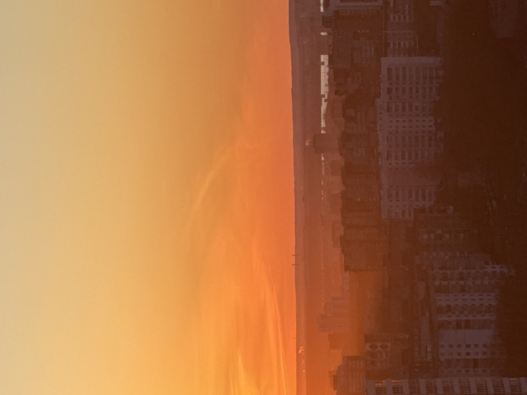 The image shows a cityscape in Perm, Russia during a sunset. The sky transitions from a light yellow to a vibrant orange. On the right side of the image are multiple buildings.