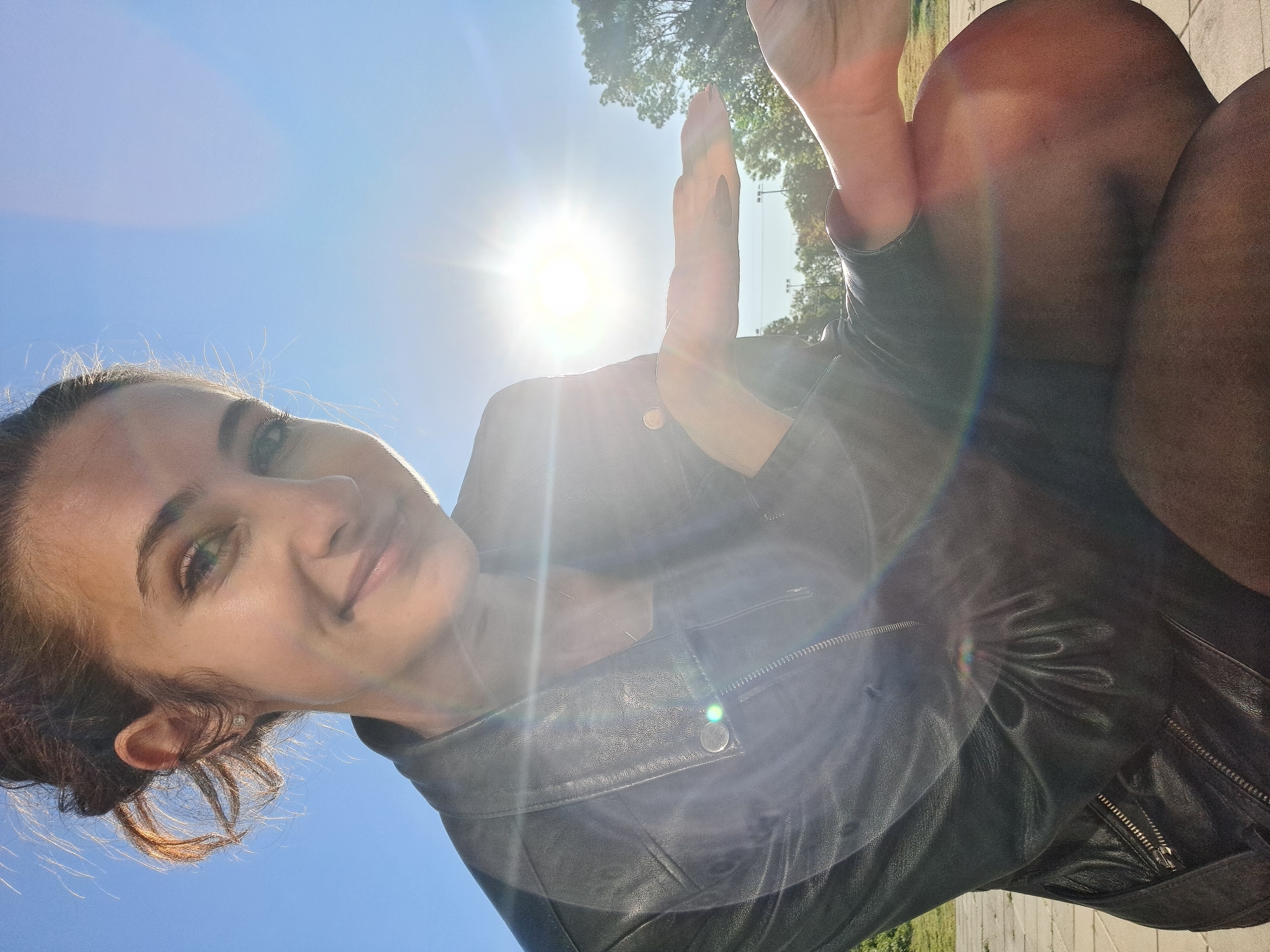 A low-angle, close-up shot depicts a woman with her legs raised, obscuring much of the background beyond a clear blue sky and a bright sun. She has light skin, brown hair pulled back, and is wearing a black leather jacket. Her face is angled towards the camera with a slight smile. Her legs are bent with her feet positioned above her head, partially blocking the sunlight. The location is likely outdoors in a city, as suggested by the presence of a paved surface and trees visible in the background, but the specific city and its exact location are unknown.