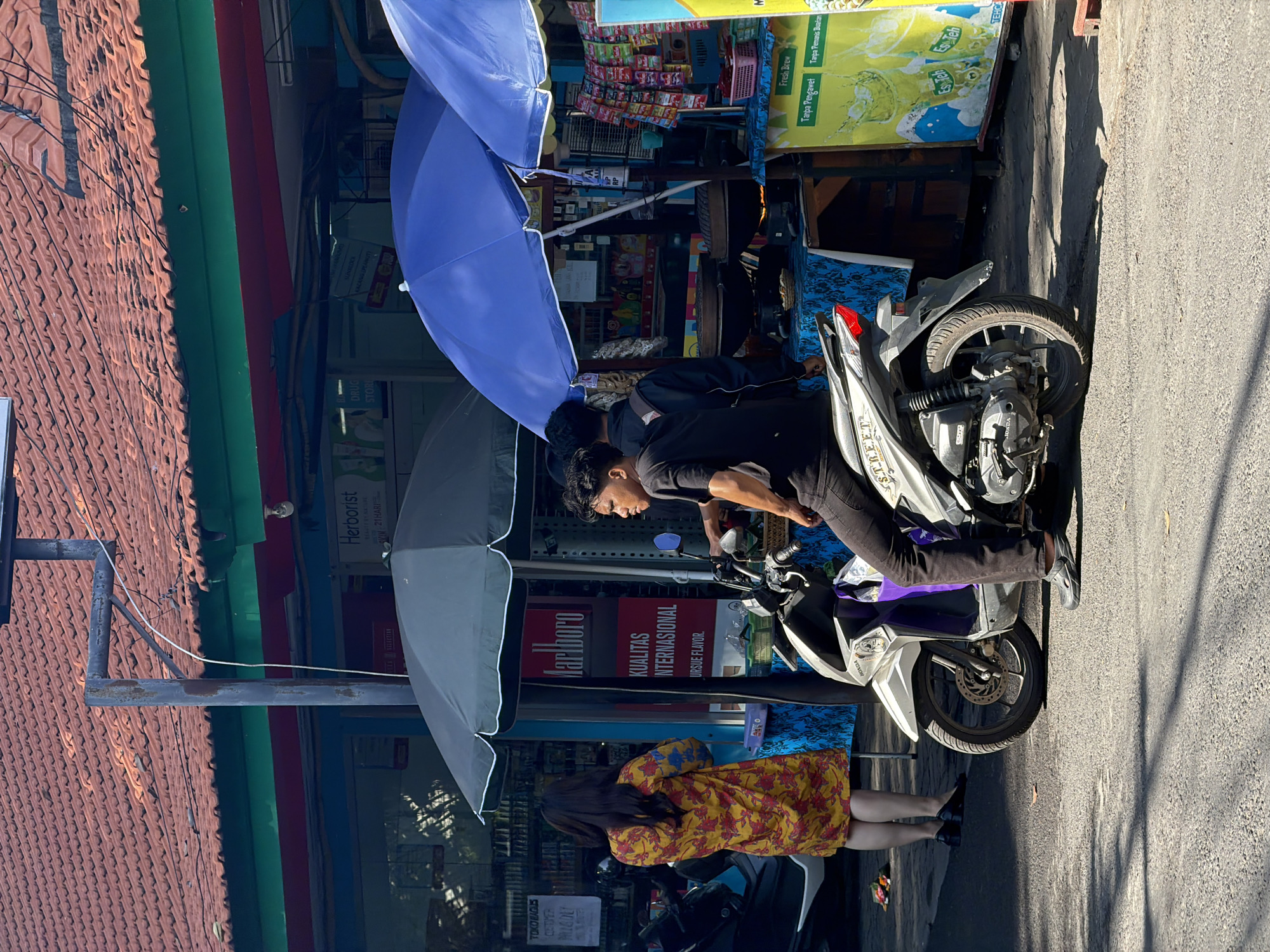 The image shows a street scene in Denpasar, Indonesia. A young man is sitting on a white motorcycle, looking to the right. He is wearing a black t-shirt and dark pants. Behind him, a woman in a yellow and red floral dress is standing and looking at a store display. The store appears to be a small convenience store or a street vendor stall, with umbrellas providing shade. Various products are visible on shelves and hanging displays, including cigarettes and packaged food items. The storefront has signs in Indonesian, and one prominently displays the Marlboro logo. The street is paved, and there are shadows cast by the sun. In the background, a building with a red tiled roof is visible. The overall atmosphere suggests a typical day in a bustling Indonesian city.