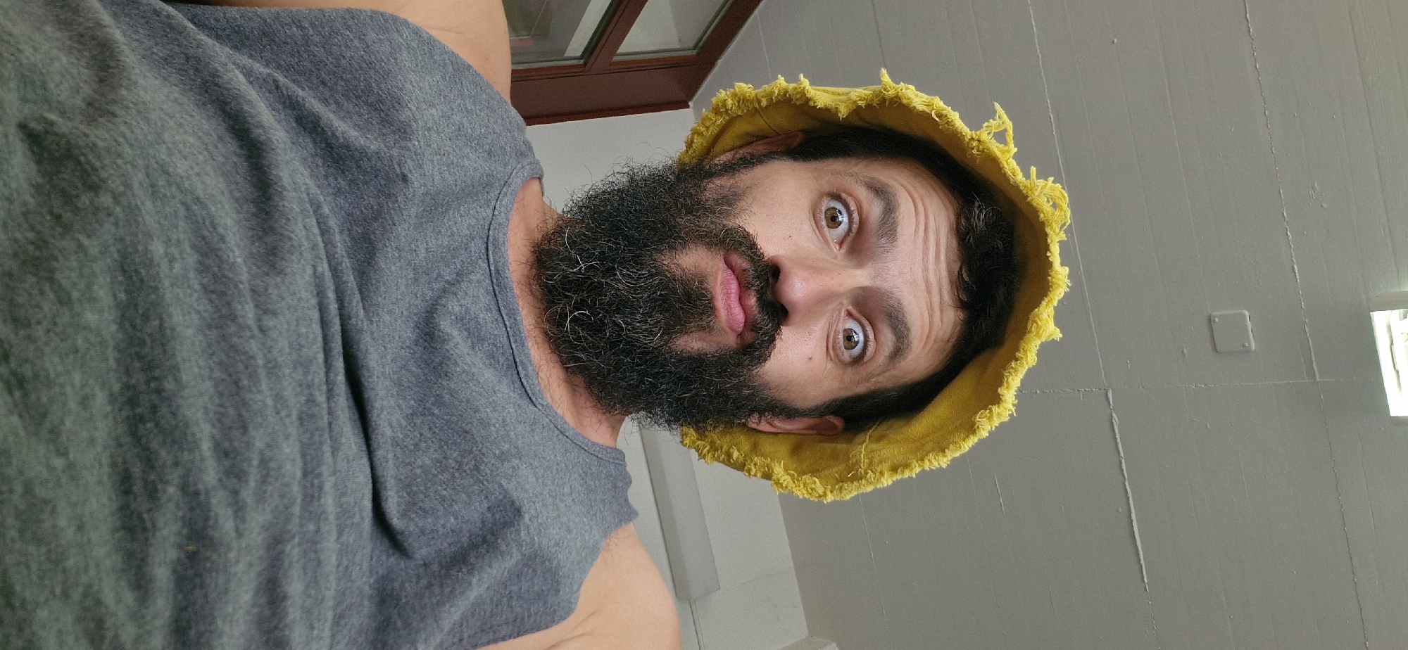 A man wearing a yellow bucket hat and a grey tank top is looking directly at the camera with a surprised expression. His eyes are wide open, and his mouth is slightly pursed. He has a full beard. The background appears to be an indoor setting with a white ceiling and a light fixture. The image was taken in Uetendorf, Switzerland.