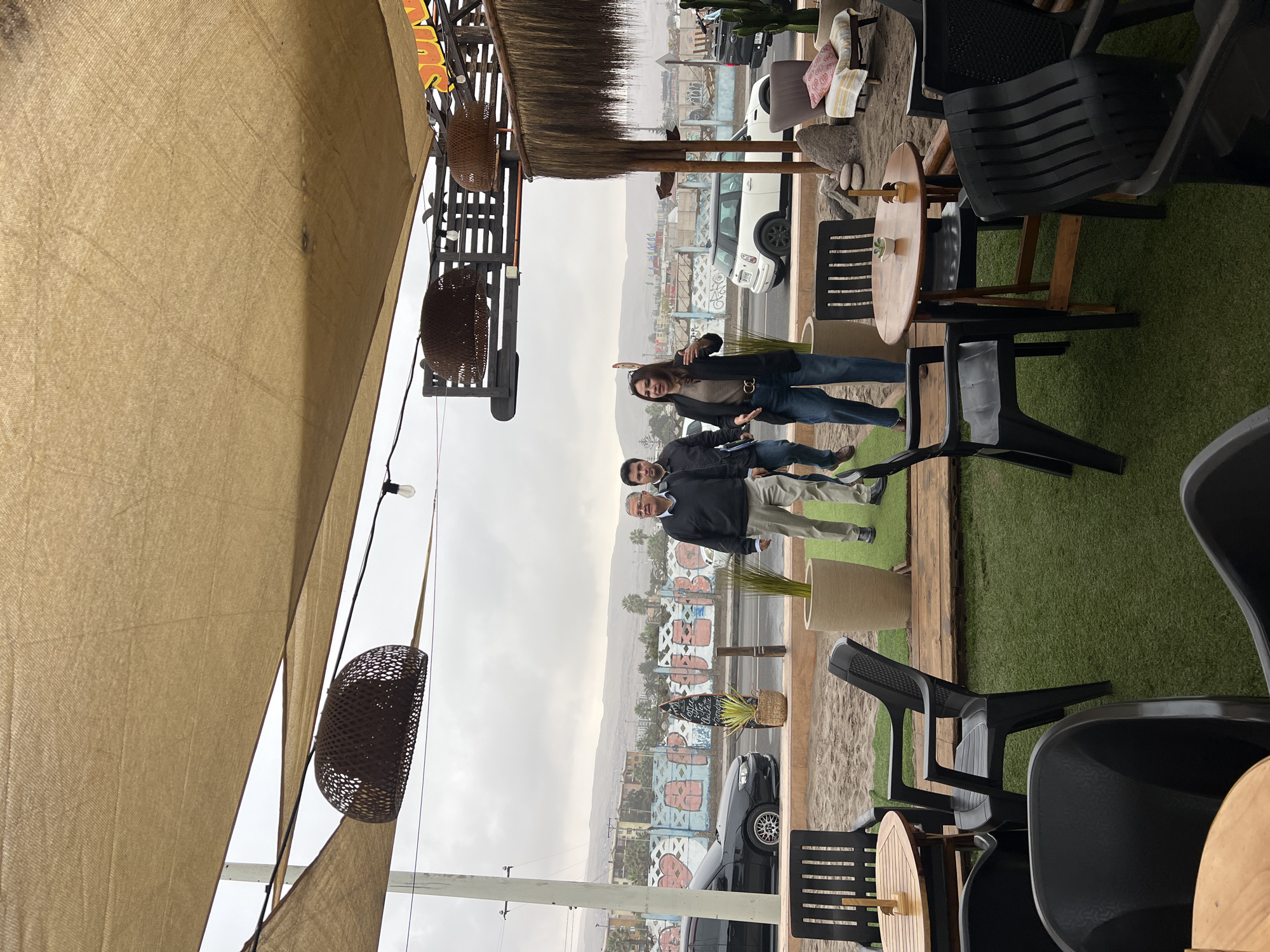 The image shows an outdoor seating area, possibly a cafe or restaurant, with tables and chairs on artificial turf. In the background, there is a street with cars and a mural on a wall. Beyond the street, there are buildings and hills, characteristic of Arica, Chile. Three people are standing in the middle ground, appearing to be engaged in conversation. The overall atmosphere suggests a casual, urban setting with a view of the city.