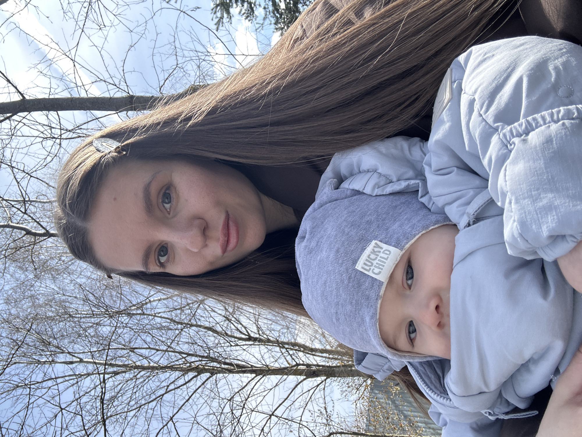 A woman with long brown hair and a baby are outdoors. The woman is wearing a hair clip and the baby is bundled in a blue hooded jacket with the words "LUCKY CHILD" on the hood. Bare trees with a blue sky background. The location is in the city of Мещёры, Russia.
