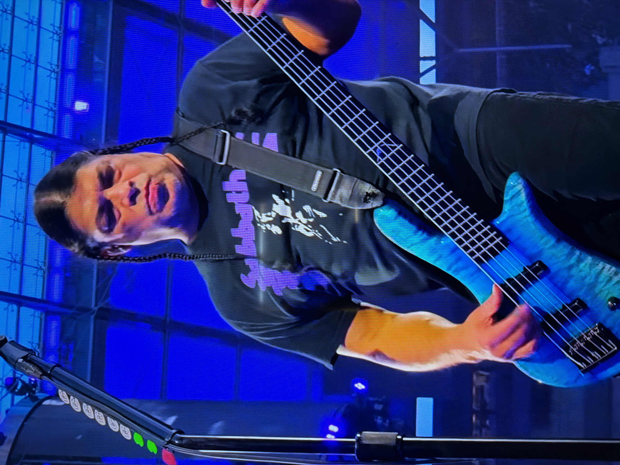 In Sacramento, California, a man with dark hair in braids is playing a blue bass guitar. He is wearing a black t-shirt with a purple graphic of a band logo. The man's left hand is on the neck of the guitar, and his right hand is strumming the strings. He appears to be performing on a stage, with a large blue backdrop behind him. The lighting is dramatic, with spotlights illuminating the performer. A microphone stand is visible to the left of the frame, with green and white lights on it. The image is taken from a slightly low angle, looking up at the musician.