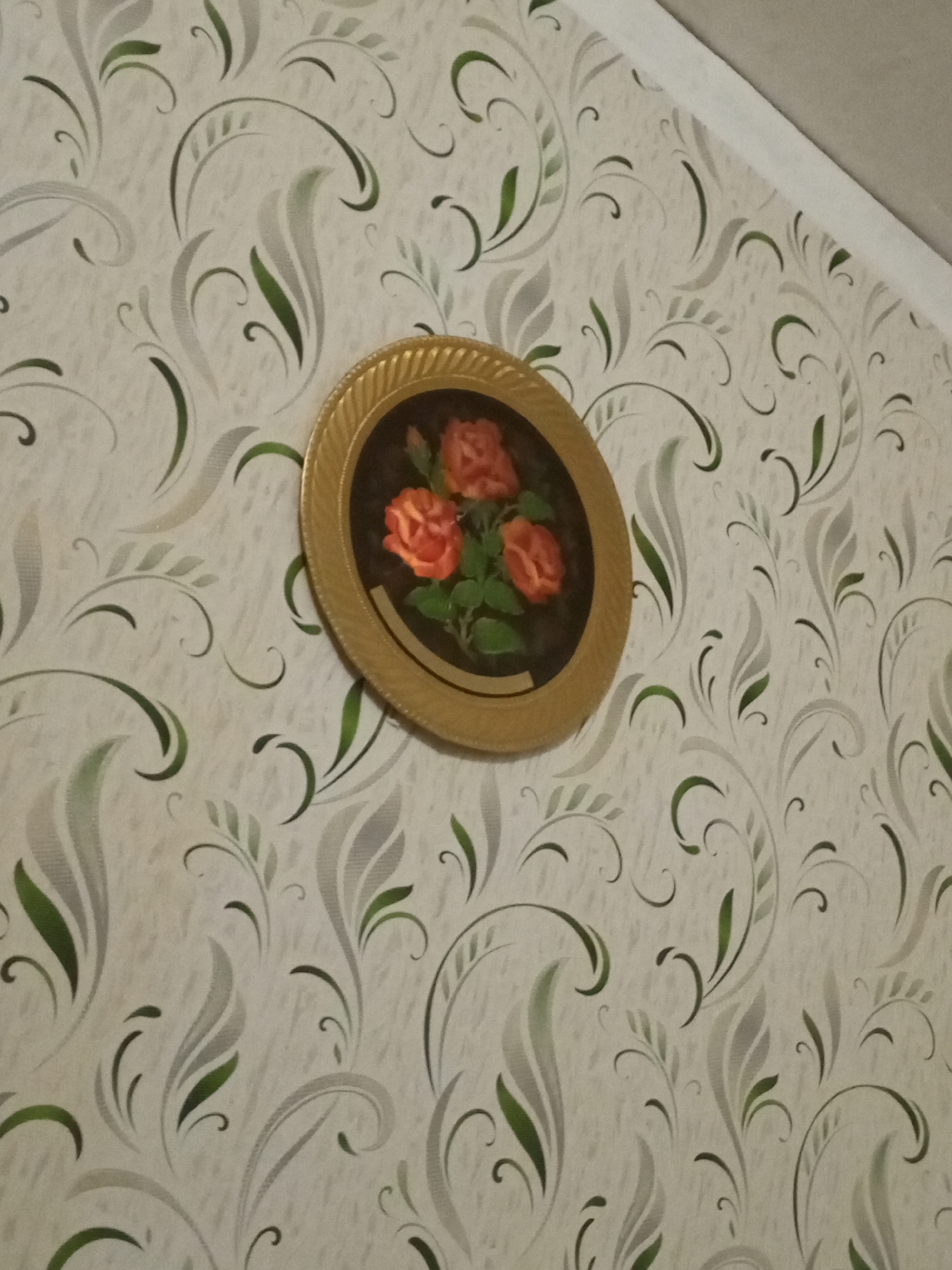 The image shows a decorative plate with a painting of three orange roses and green leaves on a patterned wallpaper background. The wallpaper has a light cream base with a swirling, floral design in shades of light gray and green. The plate is round with a textured gold-colored rim and is hanging on the wall. The scene appears to be indoors, likely in a room. The image is taken from a slightly elevated angle, looking down at the plate. The location provided, Старое Ромашкино, Russia, is a geographical reference.