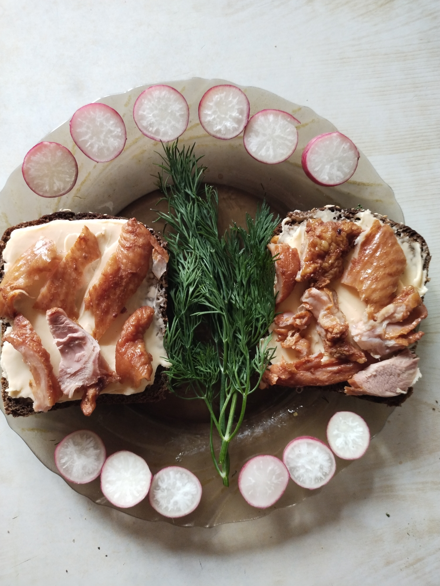 The image displays a culinary arrangement on a clear, irregularly shaped glass plate set on a light-colored, textured surface, likely a wooden tabletop, in Horishni Plavni, Ukraine. Two open-faced sandwiches, prepared on dark brown bread, are positioned centrally on the plate. Each slice of bread is spread with a pale, creamy substance, likely butter or a soft cheese. The sandwiches are topped with irregular pieces of what appears to be roasted or smoked poultry, exhibiting a range of golden-brown to reddish-brown hues, with some pieces showing lighter internal meat and visible skin. Between the two sandwiches, a fresh bundle of vibrant green dill sprigs serves as a garnish. Surrounding the upper and lower curved edges of the plate are uniformly sliced radishes, distinguished by their white centers and distinct pink-red rims, arranged in semicircular patterns. The lighting suggests an overhead or ambient source, creating minimal shadows. No human presence or interaction is visible.