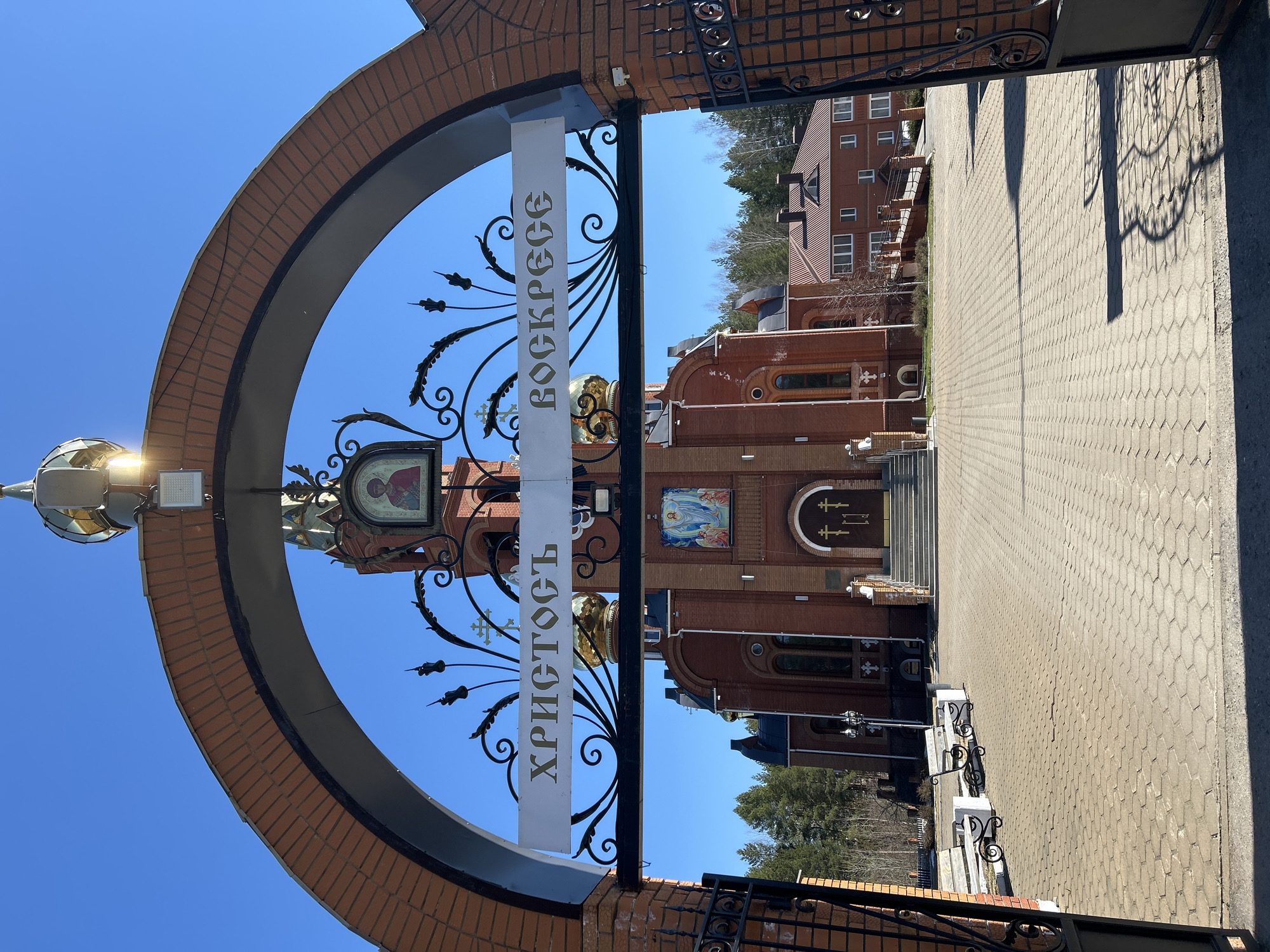The image depicts an external view of what appears to be a Russian Orthodox church in Chaikovsky, Russia. The church is made of red brick. The architecture includes domes with crosses. In front of the church stands a large, ornate wrought-iron archway. The archway has a sign displaying the words "ХРИСТОСЪ ВОСКРЕСЕ" (Christ is Risen), indicating the religious nature of the site. The ground leading up to the church is paved with interlocking stones. In the background, there is a multi-story building, also of brick. The sky is blue. No people are visible in the image.