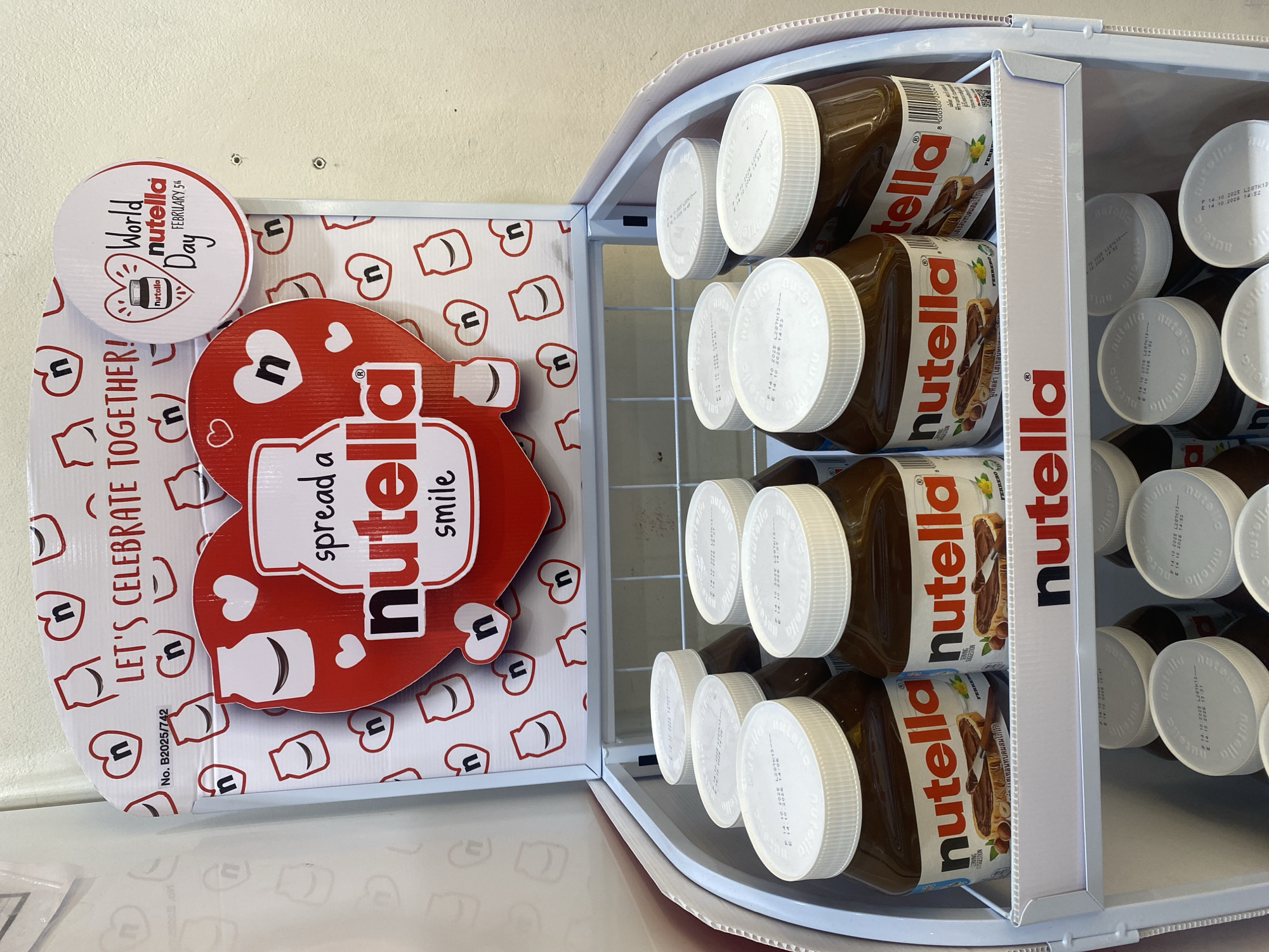 An indoor retail display features numerous large jars of Nutella spread within a white, open-top cooler or shelving unit. The jars are stacked horizontally, with their white, embossed "nutella" lids facing upwards and outwards. Each jar's label clearly displays the "nutella" logo, images of hazelnuts and bread, and text in Thai indicating "Hazelnut paste mixed with cocoa". Visible expiry dates on the lids include "E 14 10 2026".
On the left side of the display, a bright red and white promotional cutout sign is prominent. The main part is heart-shaped and features a Nutella jar graphic, with the slogan "spread a Nutella smile". Above this, a circular sign proclaims "World Nutella Day FEBRUARY 5th". Another part of the display board encourages with "LET'S CELEBRATE TOGETHER!". The background of the display boards is patterned with small heart outlines and simplified Nutella jar shapes, along with the letter 'n'. A small product number, "No. B2025/742", is also visible on the promotional material.
The setting appears to be a retail environment, such as a supermarket or convenience store in Moo 1, Thailand, designed to promote Nutella for a specific commemorative day. No people are visible, and the scene is brightly lit by ambient indoor lighting.