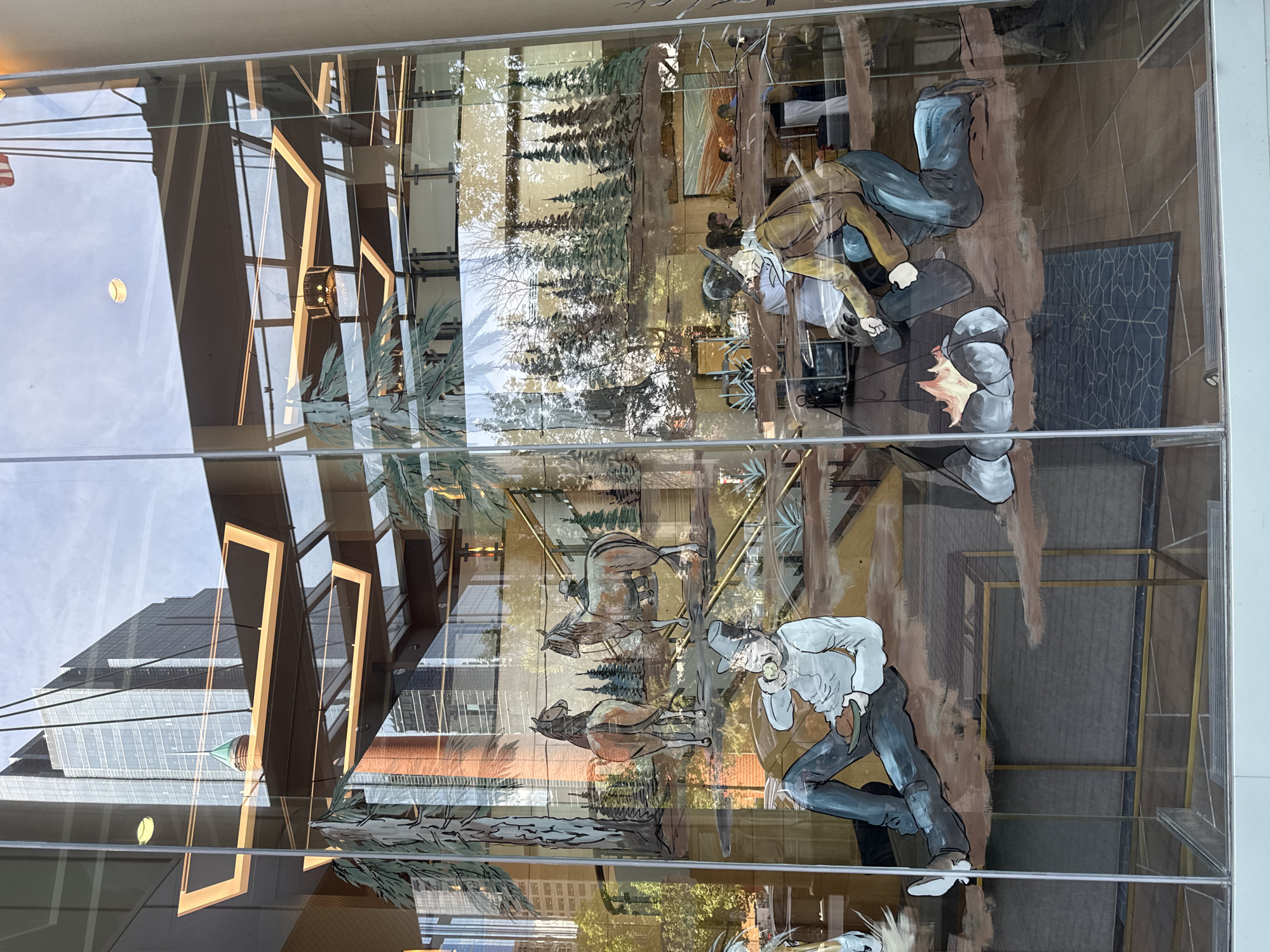 This image shows a glass window with a painted mural depicting a Western-themed scene. The mural features two individuals in the foreground, one sitting and eating, and the other kneeling by a campfire. In the background, there are images of horses, trees, and a mountain landscape. The reflection on the glass shows a modern city building with a pointed green spire, indicating the location is likely in a downtown area. Given the description and typical public art installations in Calgary, Canada, this mural is located at the Stephen Avenue Walk, a pedestrian mall in the downtown core.
The mural's style is illustrative, with bold outlines and flat areas of color. The foreground subject on the left is a person in a light blue shirt and dark pants, seated with their legs crossed, holding a piece of food and a drink. They are wearing a hat. To the right, another person in a brown jacket and blue pants is kneeling, tending to a campfire with visible flames.
The background of the mural displays two horses, one facing away from the viewer, and another facing towards the right. Behind the horses, there is a stylized depiction of pine trees, suggesting a wilderness setting.
The reflections on the glass are significant. The most prominent reflection is of a tall, modern skyscraper, characterized by its glass facade and a distinctive pointed green spire at its peak. This architectural style is consistent with buildings found in the financial district of Calgary. Other reflections include sky, other buildings, and interior elements of the building where the window is located, such as ceiling lights and structural beams. The mural is applied to the exterior of a building, viewed from the street.