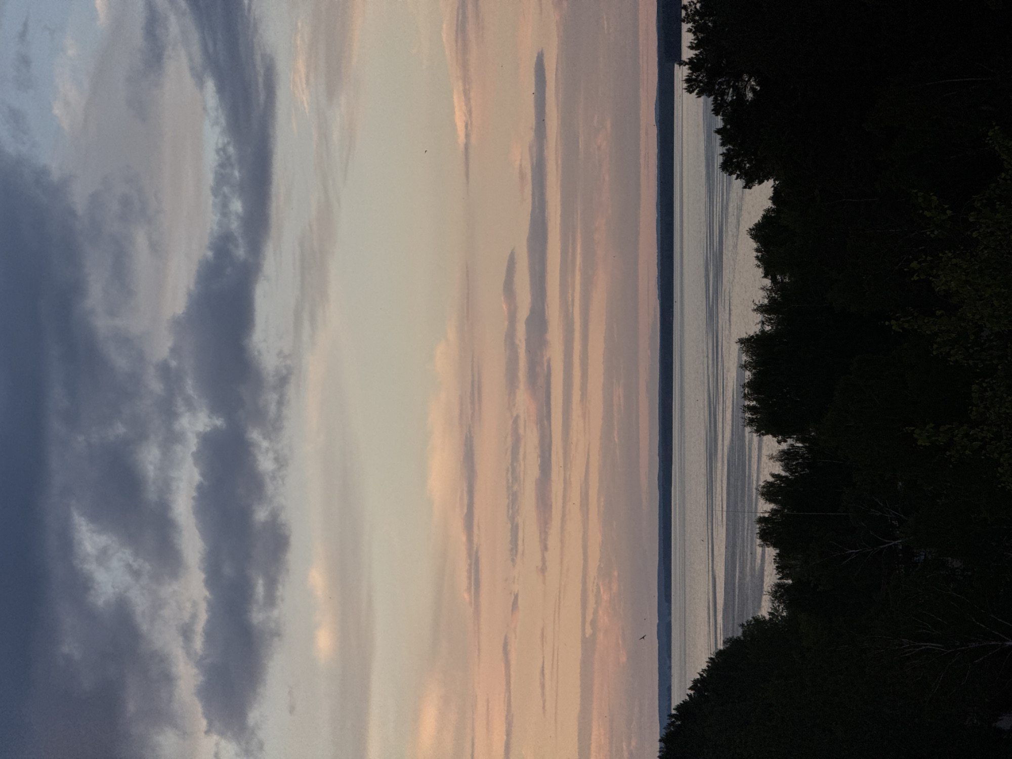 The image depicts a sunset scene over a body of water, likely a river or lake. The sky is filled with varied cloud formations; gray and blue clouds are present on the left, transitioning to lighter pink and gray striated clouds towards the right. The water reflects the sky's colors, showing similar striations of light and darker shades. The edge of a forested area is visible along the right side of the image, appearing dark. There are no people visible in the shot. The image is taken near the city of Chaikovsky, Russia.