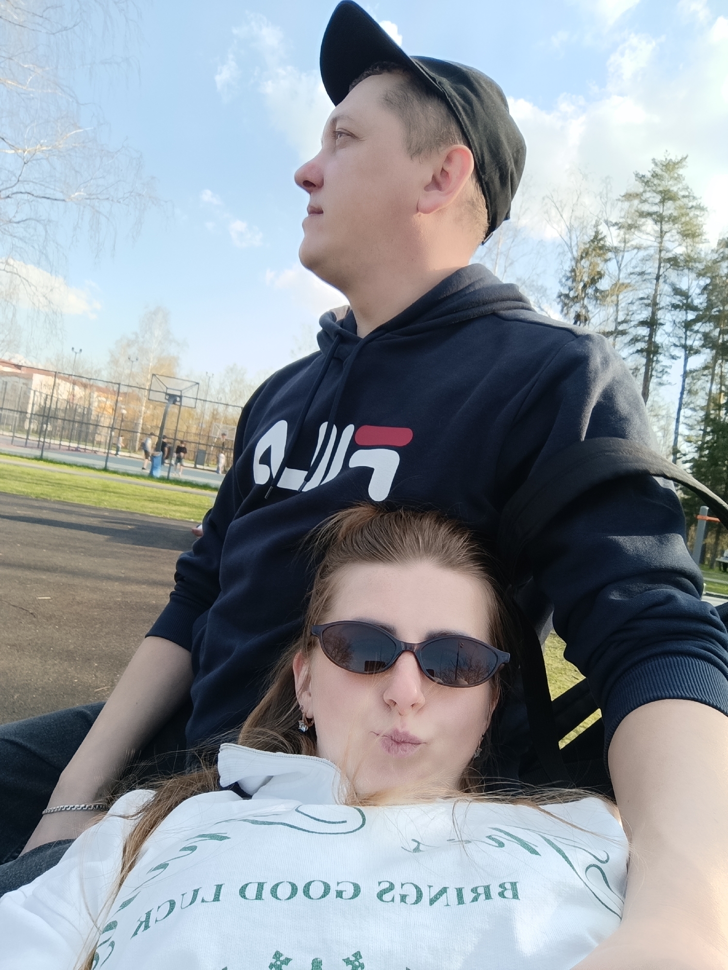 A medium shot captures a man and a woman in an outdoor setting. The man, positioned in the background, is wearing a dark blue hoodie with a large white logo on the chest and a black baseball cap. He is looking to the left of the frame. The woman, in the foreground, is wearing sunglasses and a white shirt with green text. Her head is resting against the man's chest. The background features a grassy area with a basketball court and trees, suggesting a park or recreational area. The sky is blue with scattered clouds, indicating a bright, sunny day.