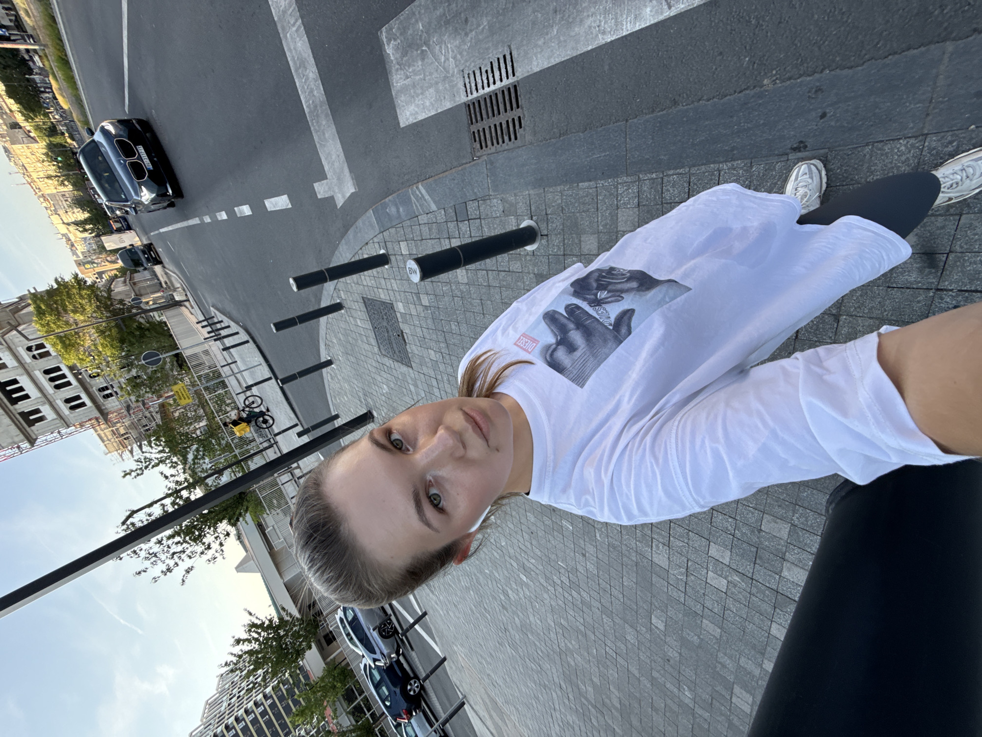 A young woman with blonde hair pulled back in a ponytail and wearing a white t-shirt with a graphic print and dark leggings stands on a paved sidewalk. She has white wireless earbuds in her ears. Behind her, cars are parked along a street, and a dark-colored sedan is driving on the road. In the background, there are buildings under construction and trees. The scene is set in a city, possibly Belgrade, Serbia, given the context. The lighting suggests it is daytime. The woman is looking towards the camera, seemingly taking a selfie.
